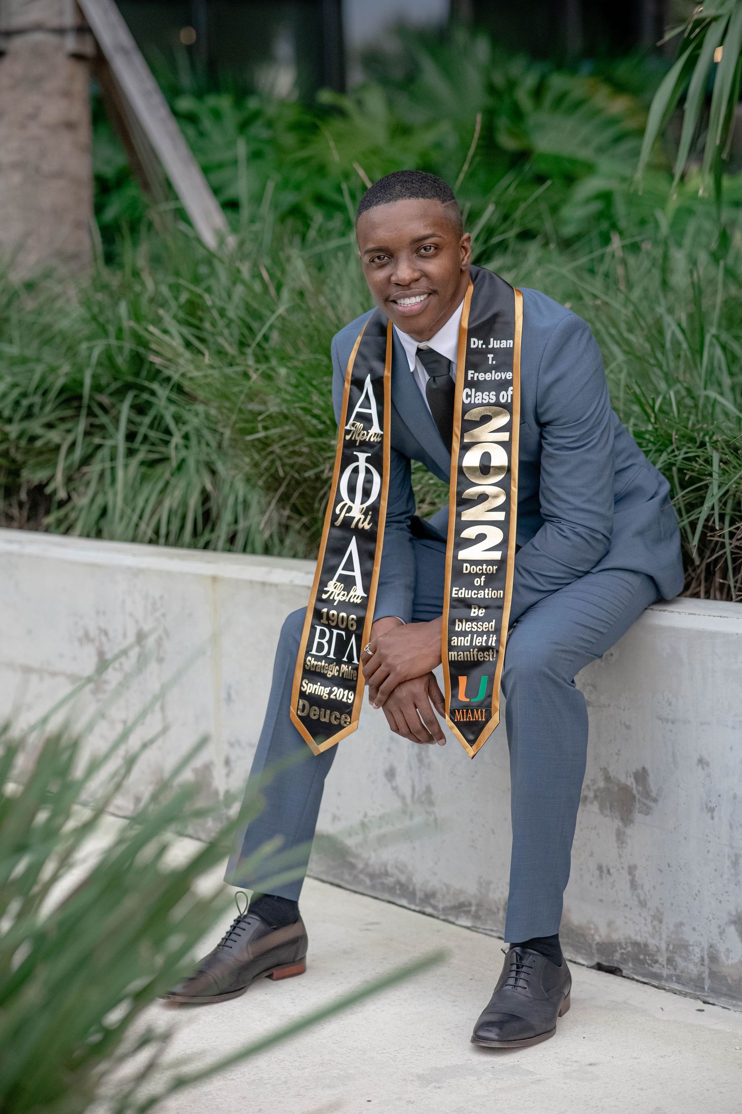 Black male University of Miami graduate sits outdoors wearing Alpha Phi Alpha graduation stole.