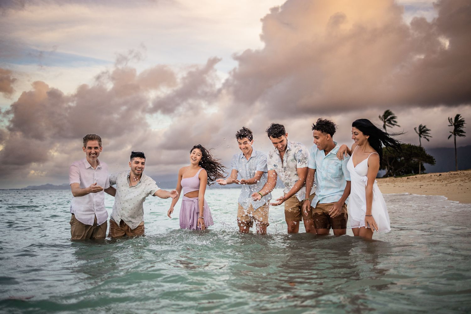 Group standing in ocean water at sunset with dramatic cloudy sky and palm trees.