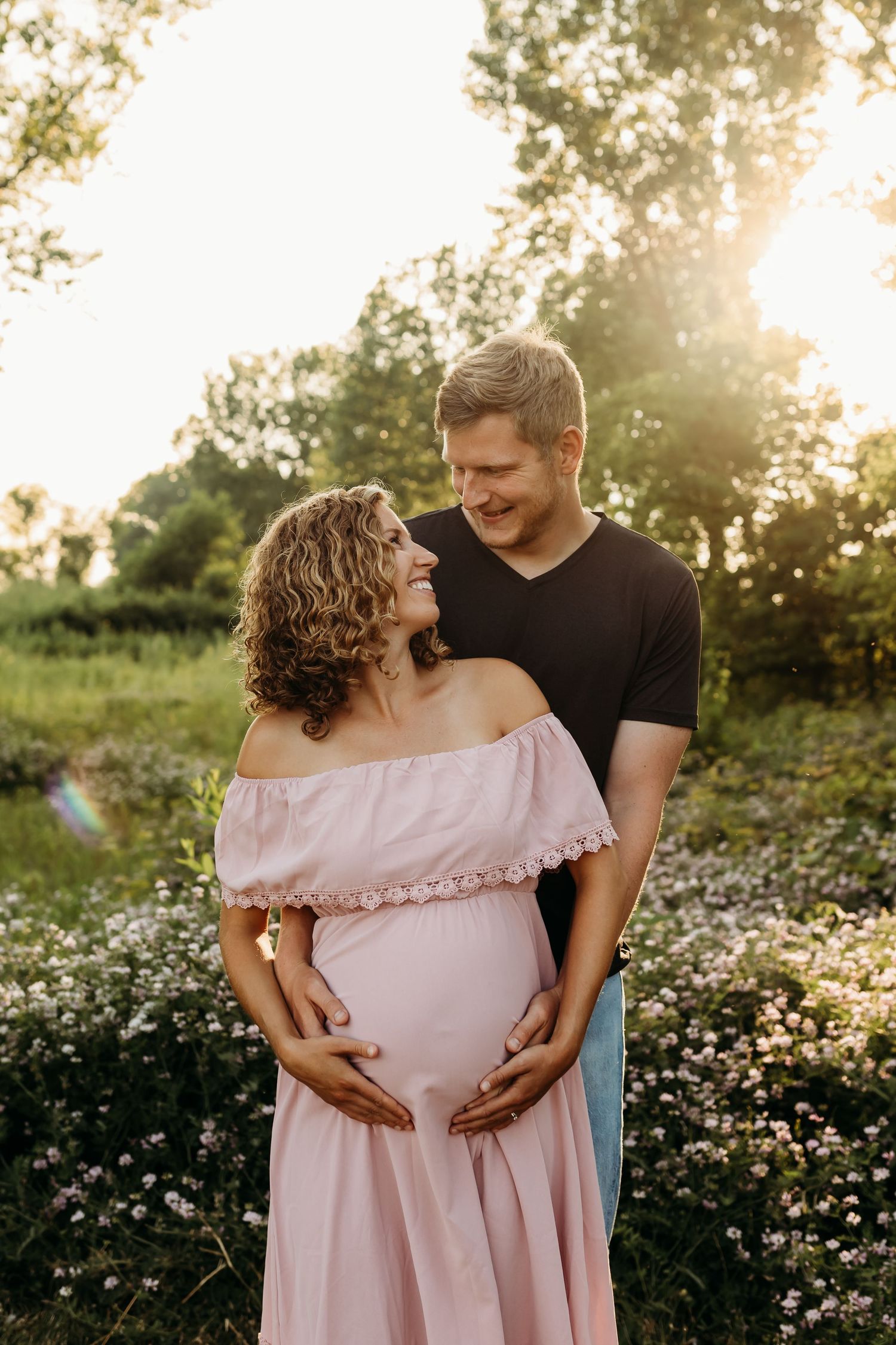 A maternity photo of a couple in a flower-filled meadow with golden sunlight streaming through trees.