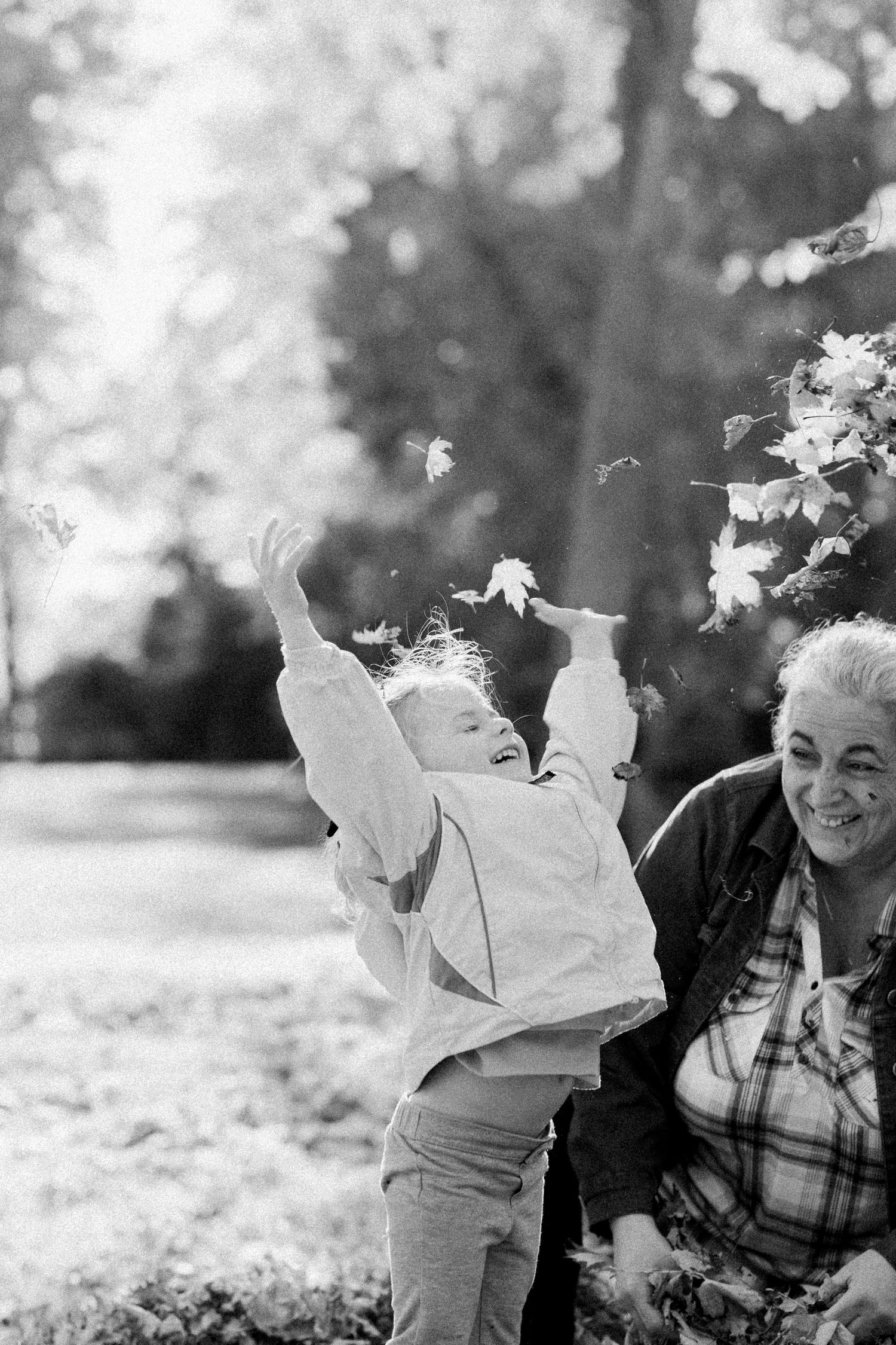 A young girl throws leaves into the air excitedly, her grandmother watching and smiling behind her. Photo by Katie Crane