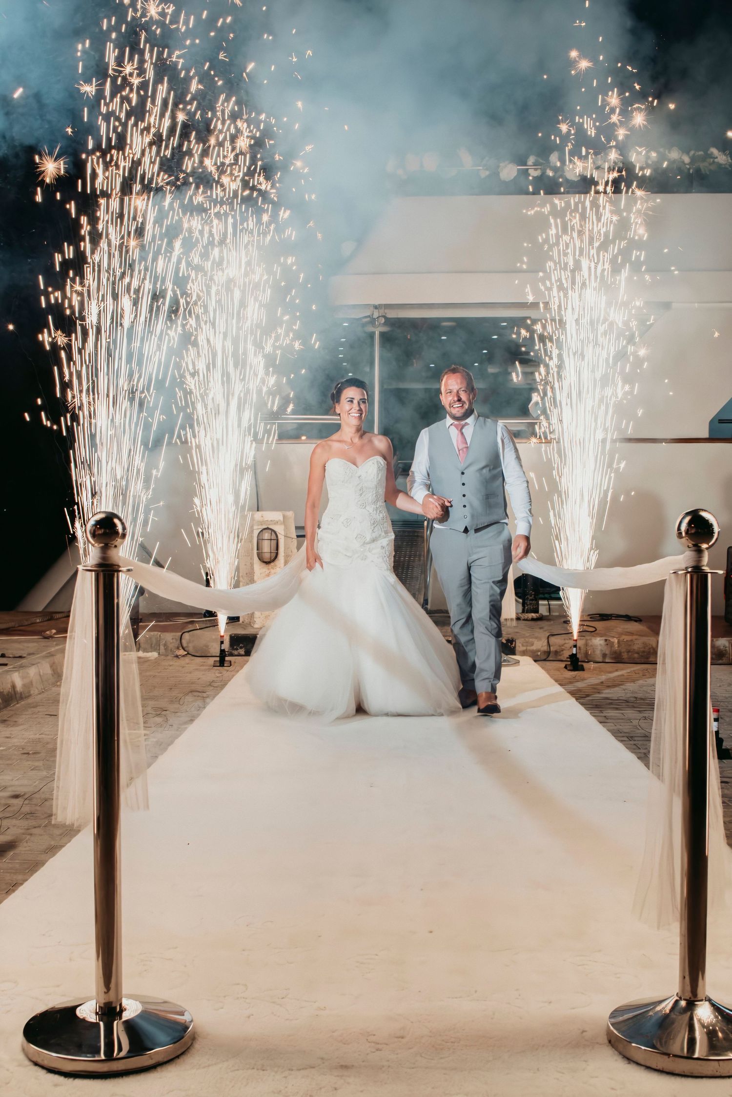 Wedding couple makes grand entrance on white carpet with sparkling firework fountains on both sides.
