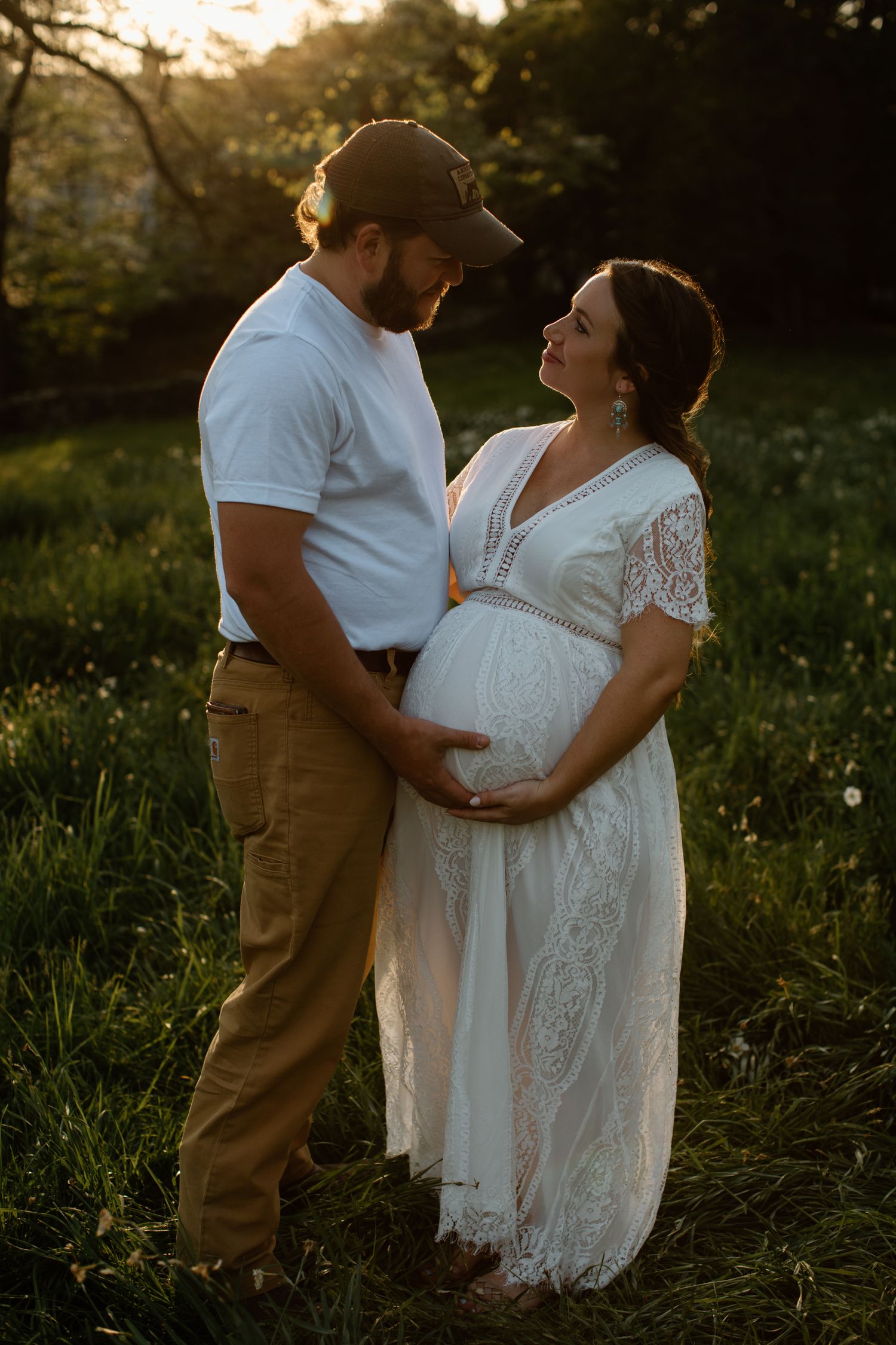 Maternity photo session in a grassy field at sunset with couple in white dress and khaki pants.
