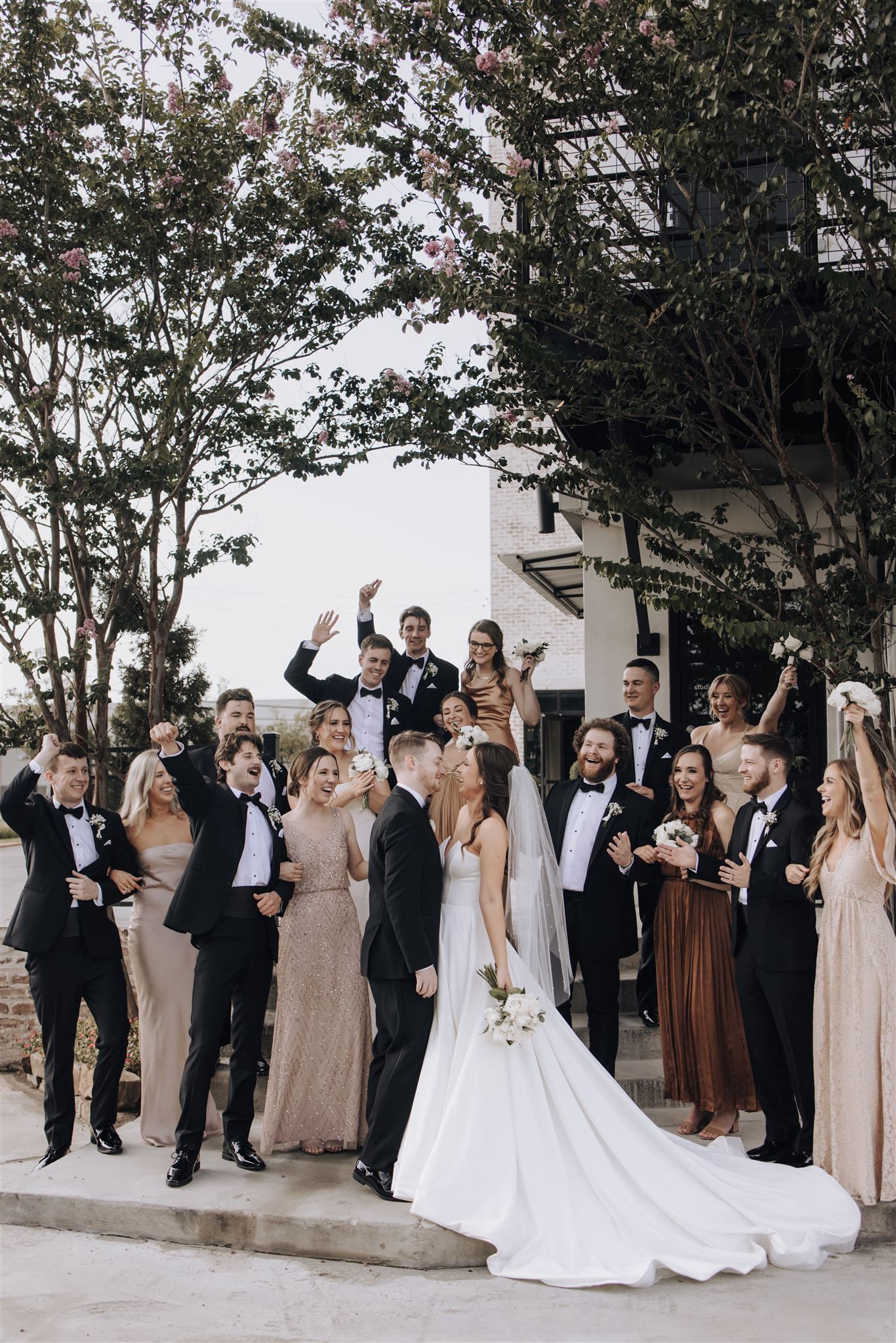 Wedding party in formal attire posing with raised arms against modern building exterior.