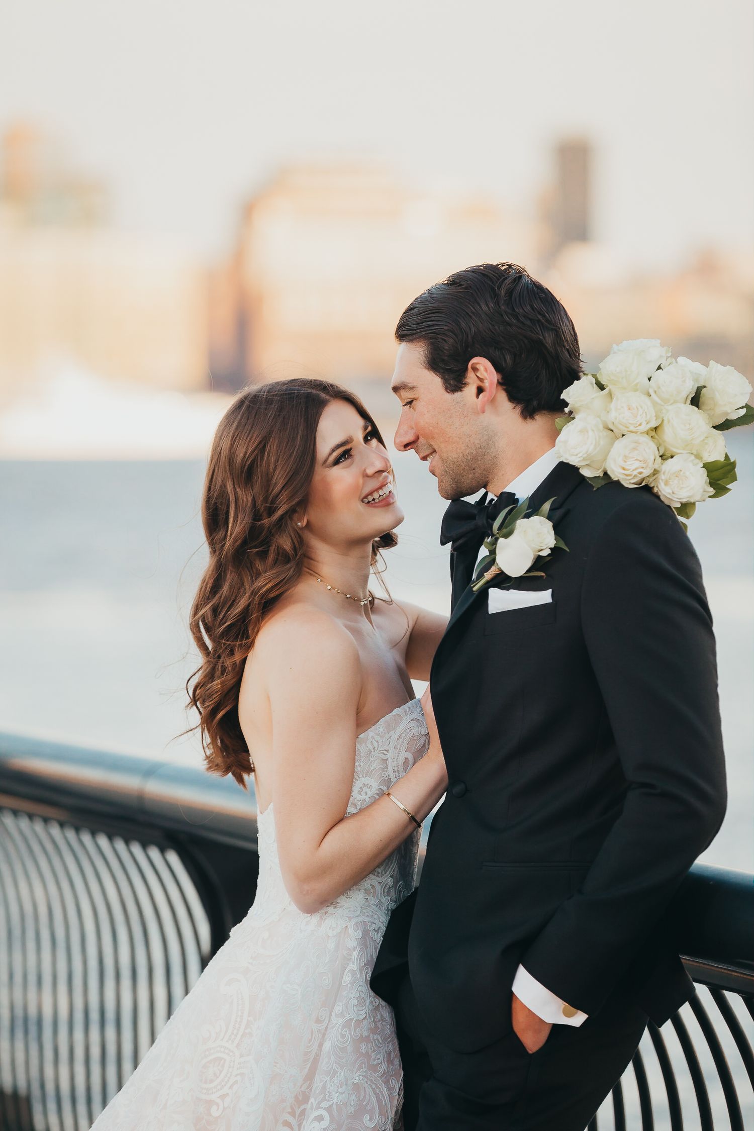Bride and groom sharing a romantic moment on a waterfront dock with city skyline at golden hour.