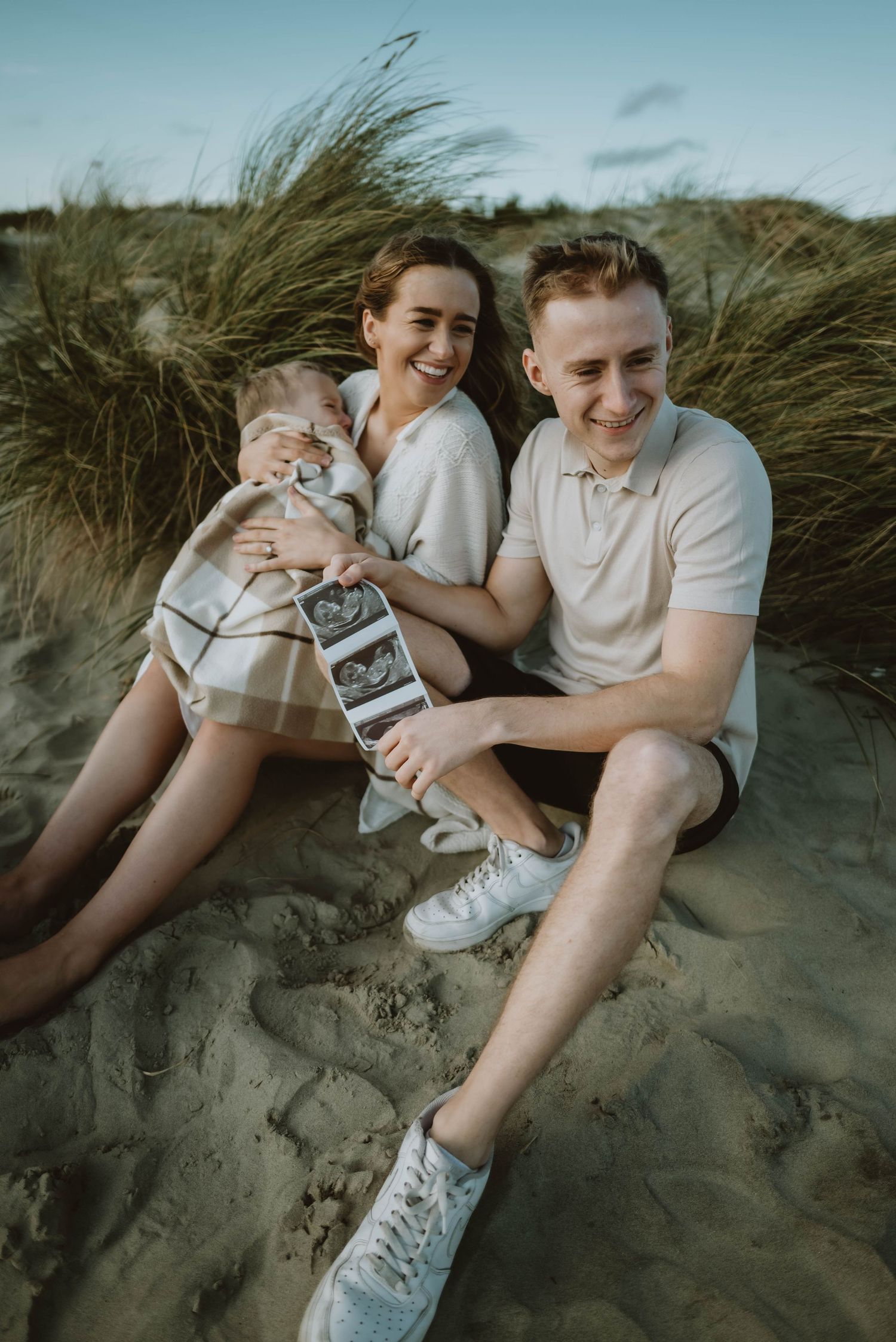 A couple sits together on sandy dunes with tall grass swaying in the background.
