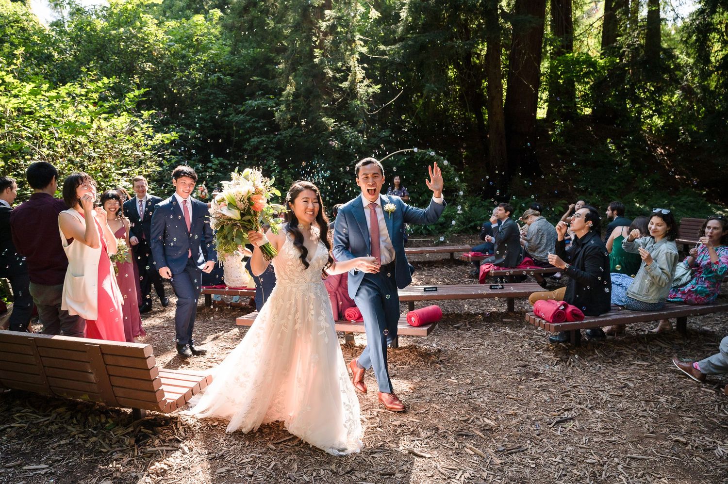Newlyweds celebrate during recessional at outdoor woodland wedding with guests on rustic benches.