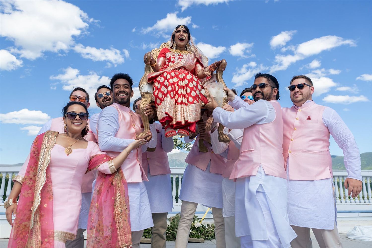 Wedding party in pink and white outfits lifting and celebrating on a sunny outdoor deck.