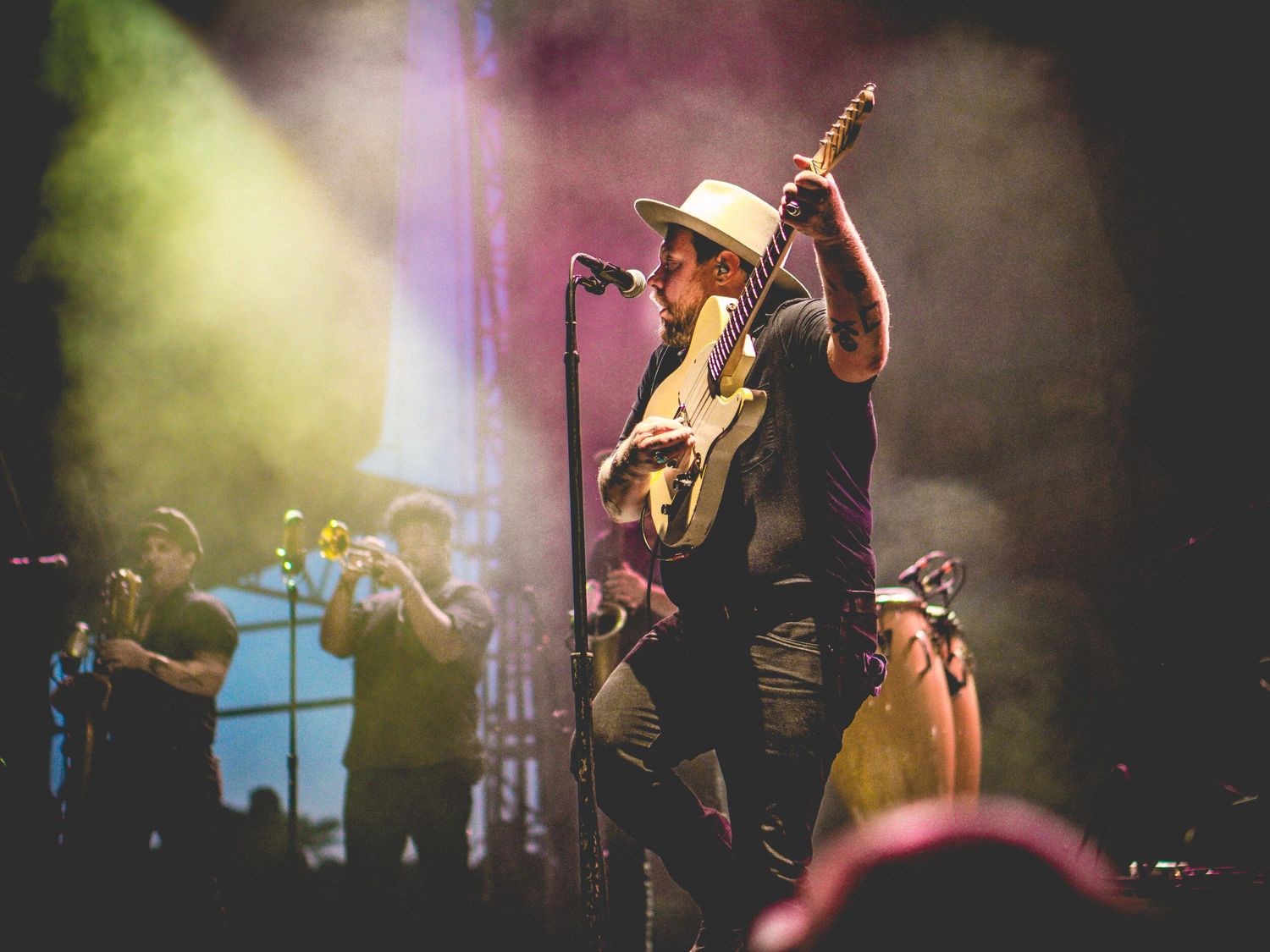 Country music performer in a cowboy hat plays guitar on stage with dramatic lighting and band members in background.
