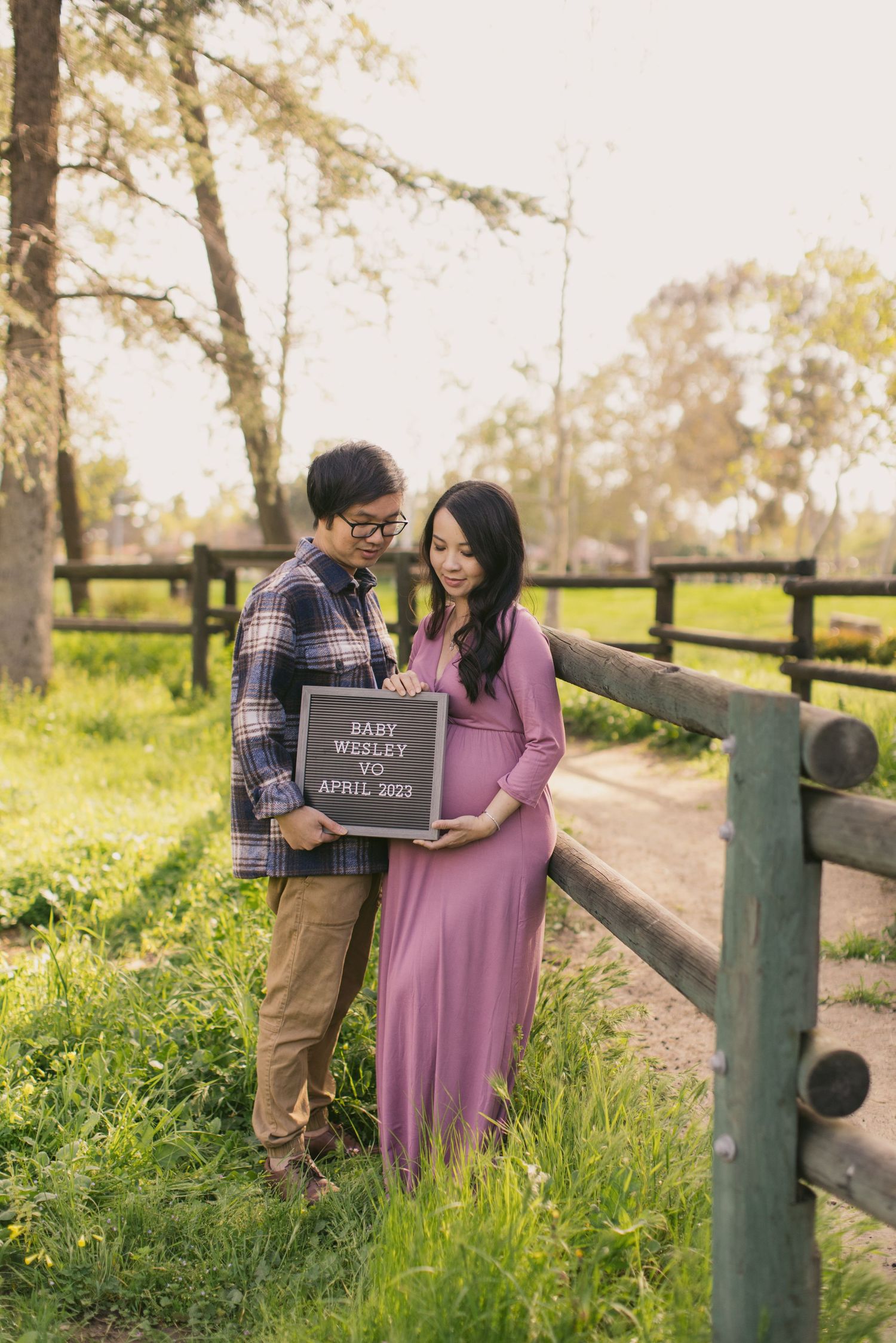 Couple holding a baby announcement sign during their maternity photoshoot in a park setting.
