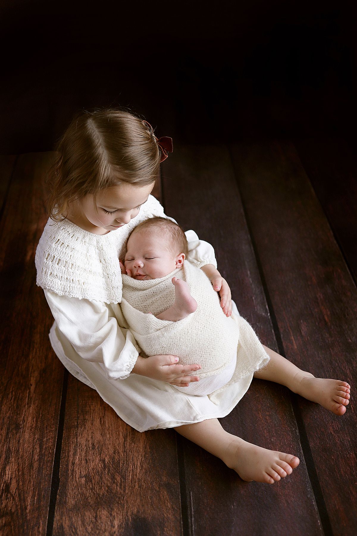 Two children in white dresses sit on dark wooden floor in tender embrace.
