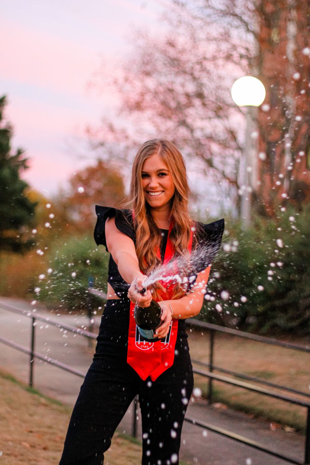 Graduate sprays champagne during sunset celebration photoshoot.
