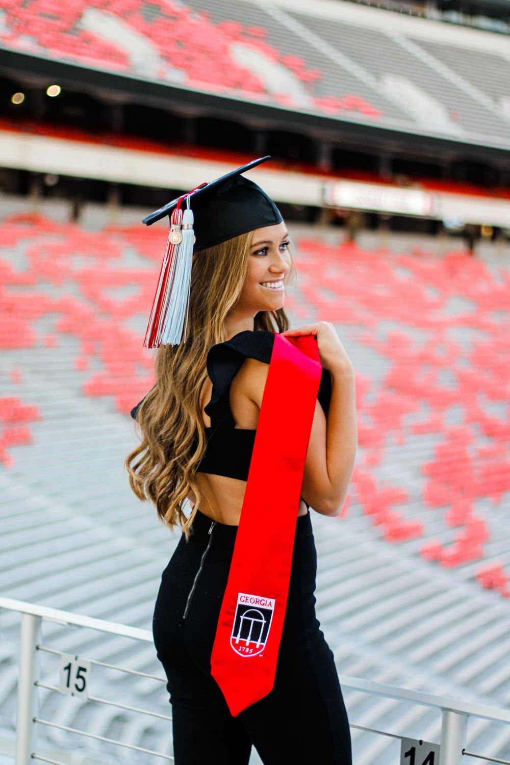 Graduate in cap and gown poses in stadium with red stole and black attire.