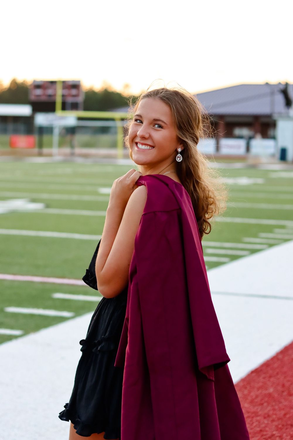 Student with burgundy cap and gown poses on football field during golden hour.