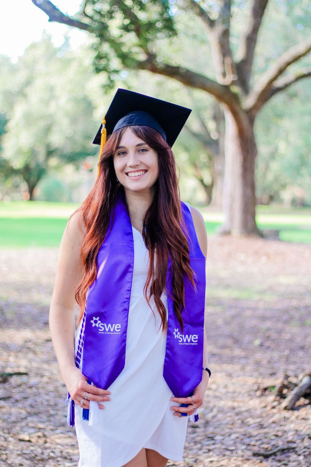 Graduate in purple SWE stole poses outdoors under tree canopy.