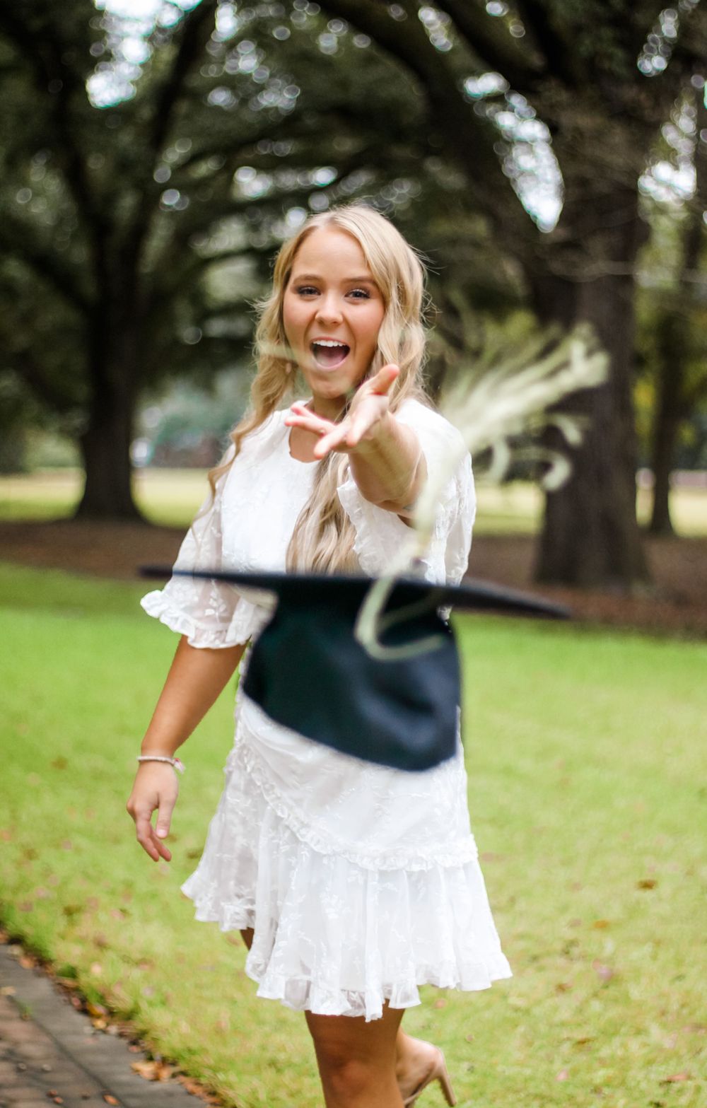 Graduate in white dress poses joyfully with cap on campus grounds.
