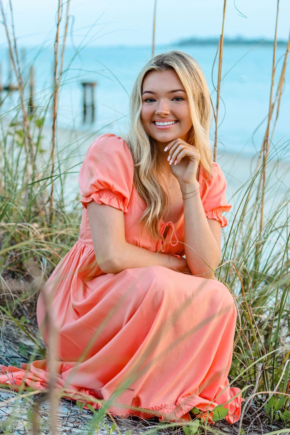 Woman in coral dress sits among beach grass with ocean view.
