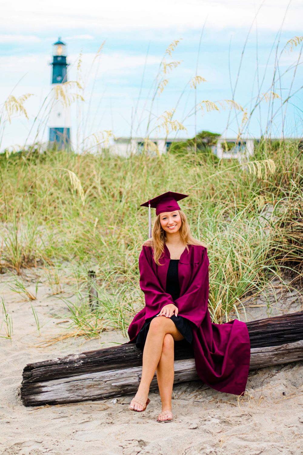 Graduate in burgundy gown poses on beach with lighthouse in background.