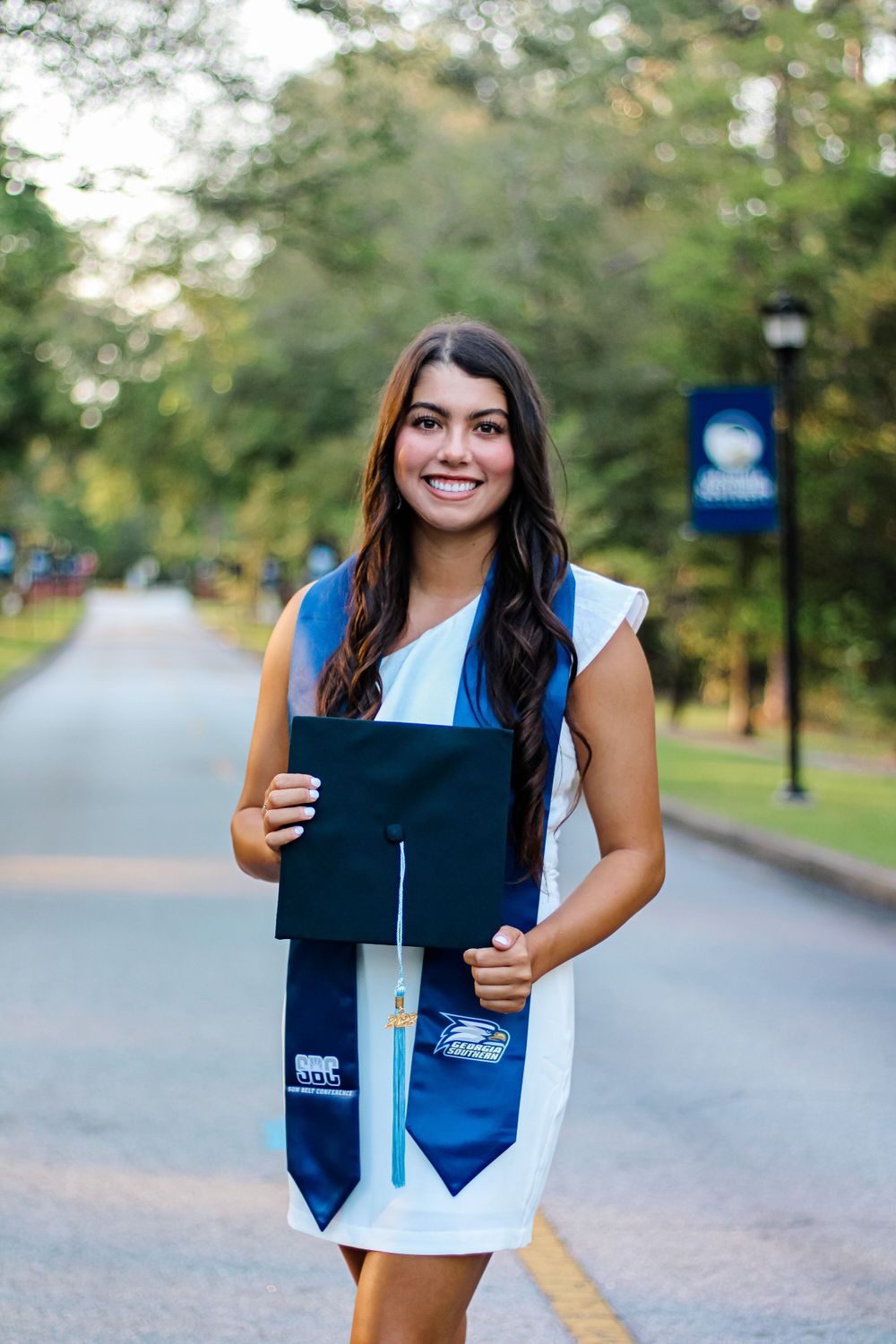 Graduate in white dress and blue stole holds cap while standing on campus pathway.