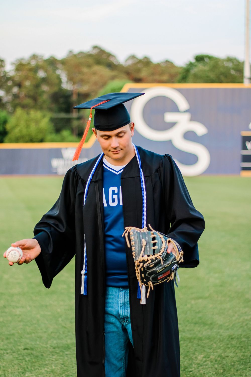 Graduate in cap and gown holds baseball glove while standing on sports field.