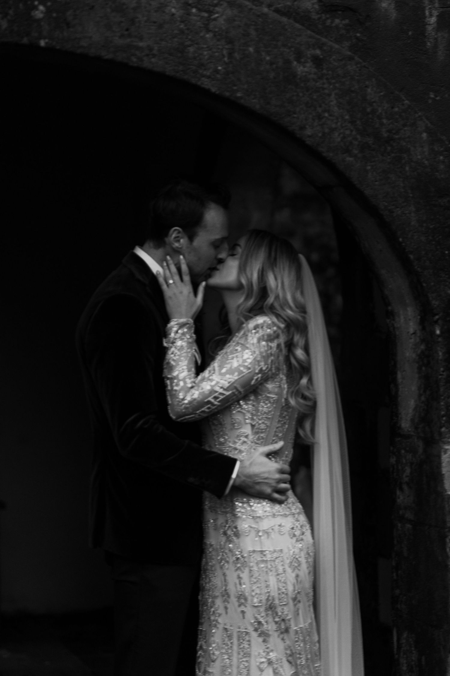 Dramatic black and white photo of couple embracing under stone archway, bride in lace gown.
