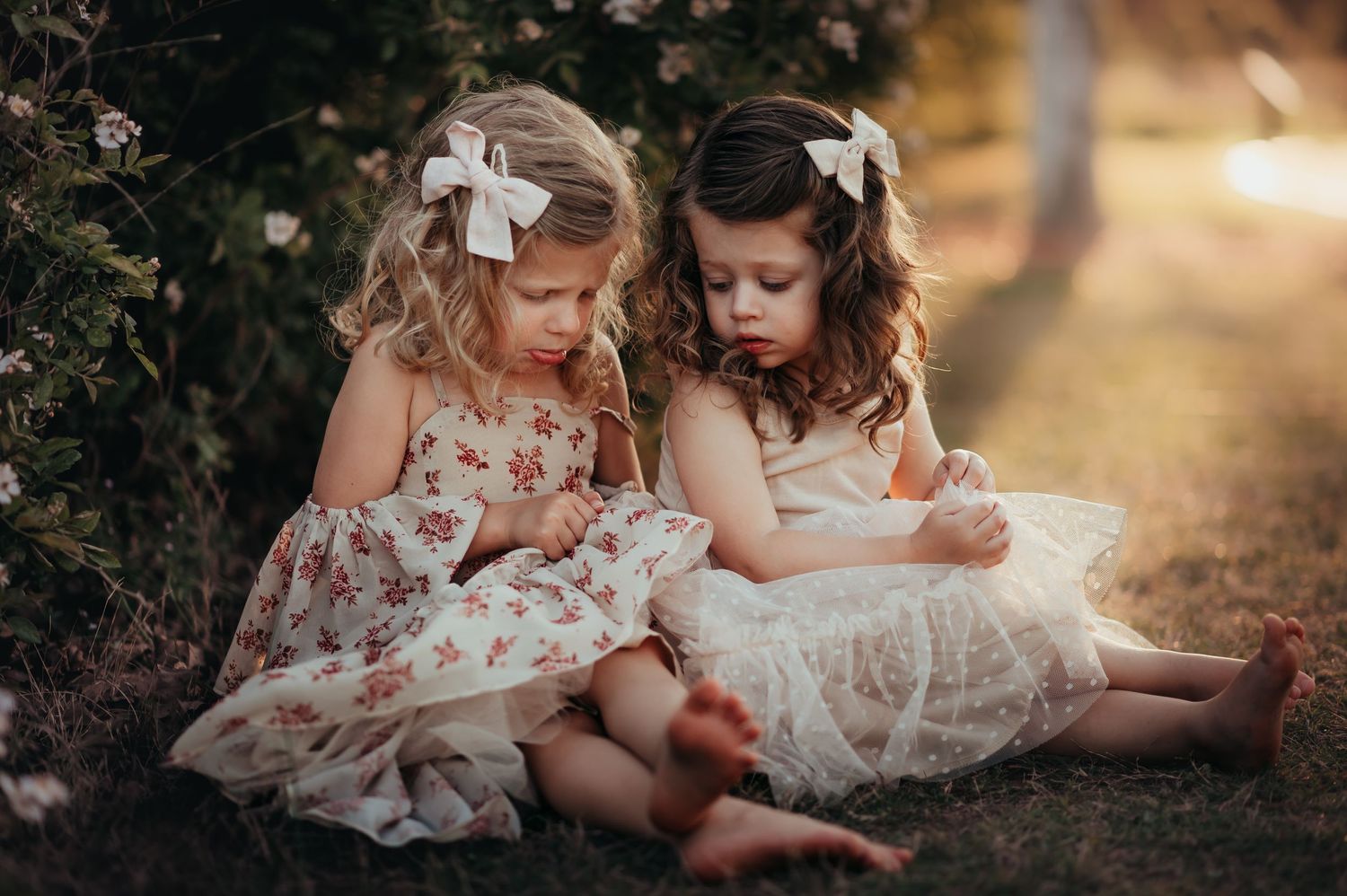 two girl cousins in pretty dresses sitting on the ground look down at their hands with sad faces.