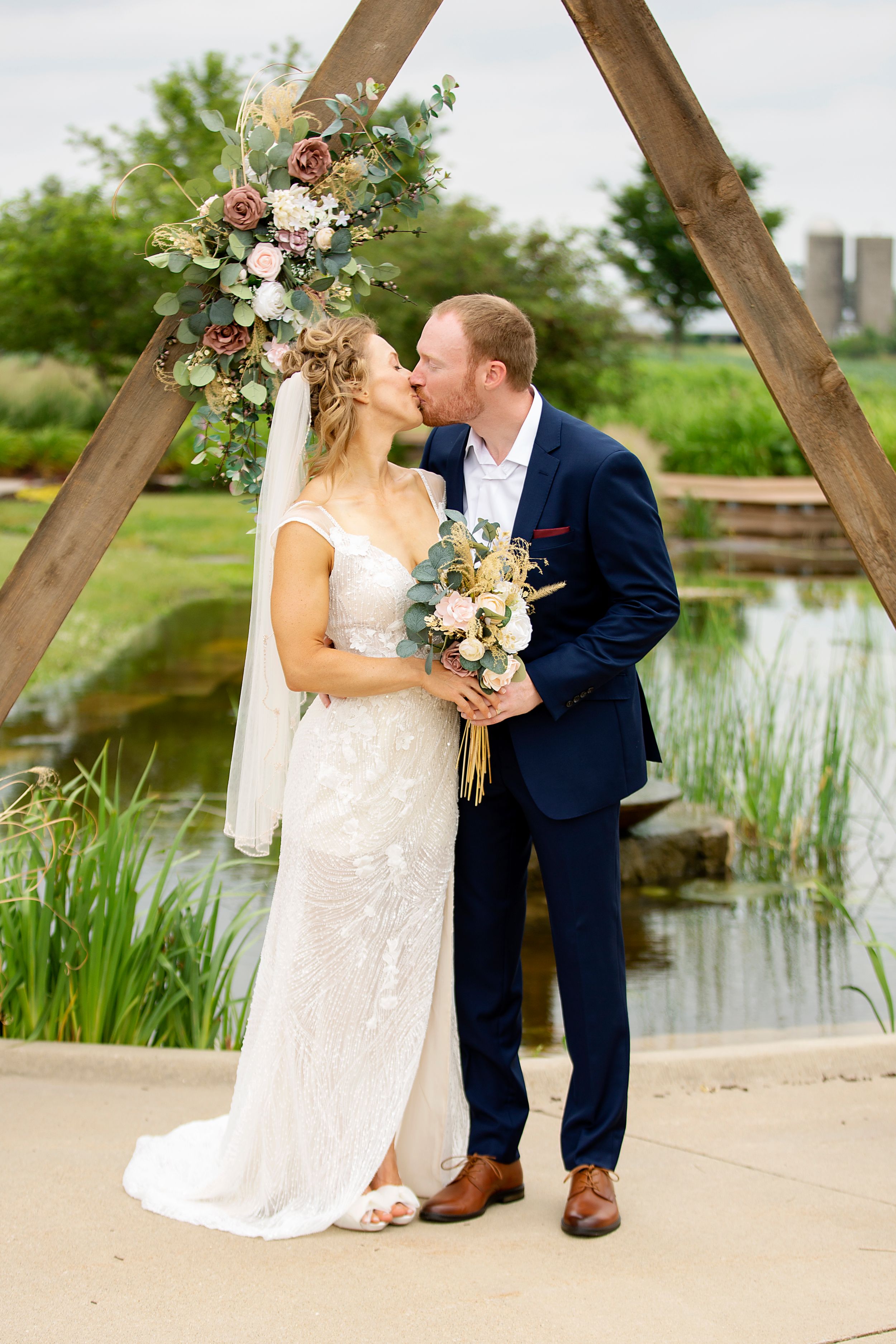 A romantic wedding portrait under a wooden arch decorated with flowers beside a pond.