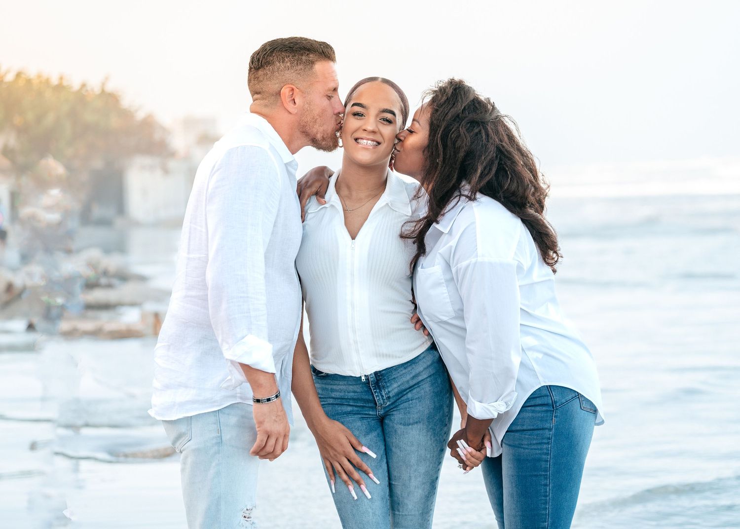 Three people share an affectionate moment on a sunny beach wearing coordinated white and denim outfits.