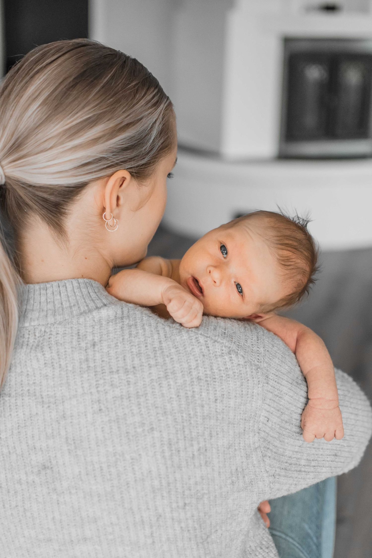 Parent in grey sweater holding sleeping baby on shoulder in kitchen.