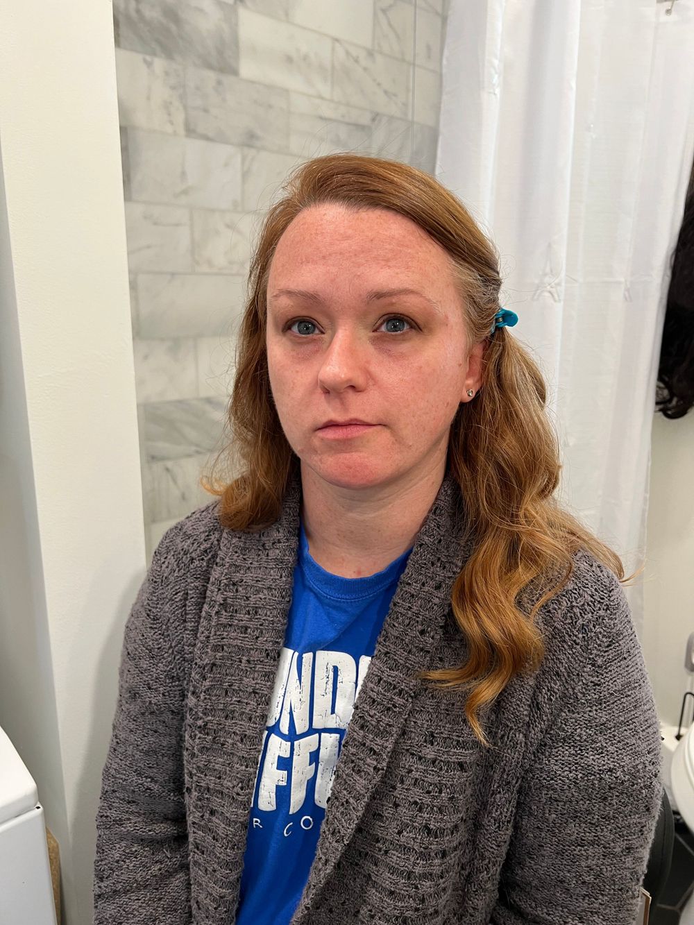 Person wearing a blue shirt and grey cardigan seated against a white wall.