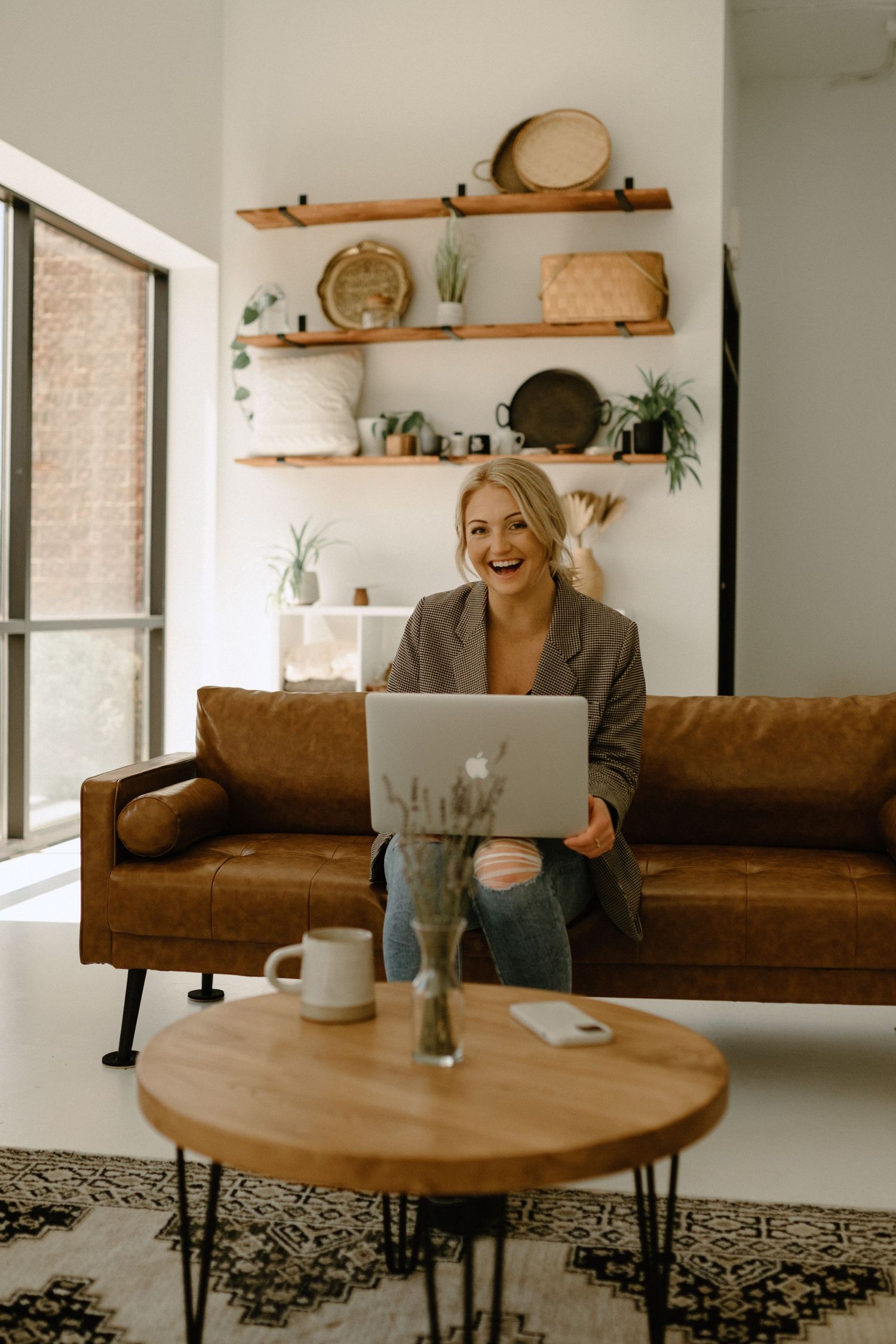 A woman works on a laptop while sitting on a brown leather couch with decorative shelving in the background.