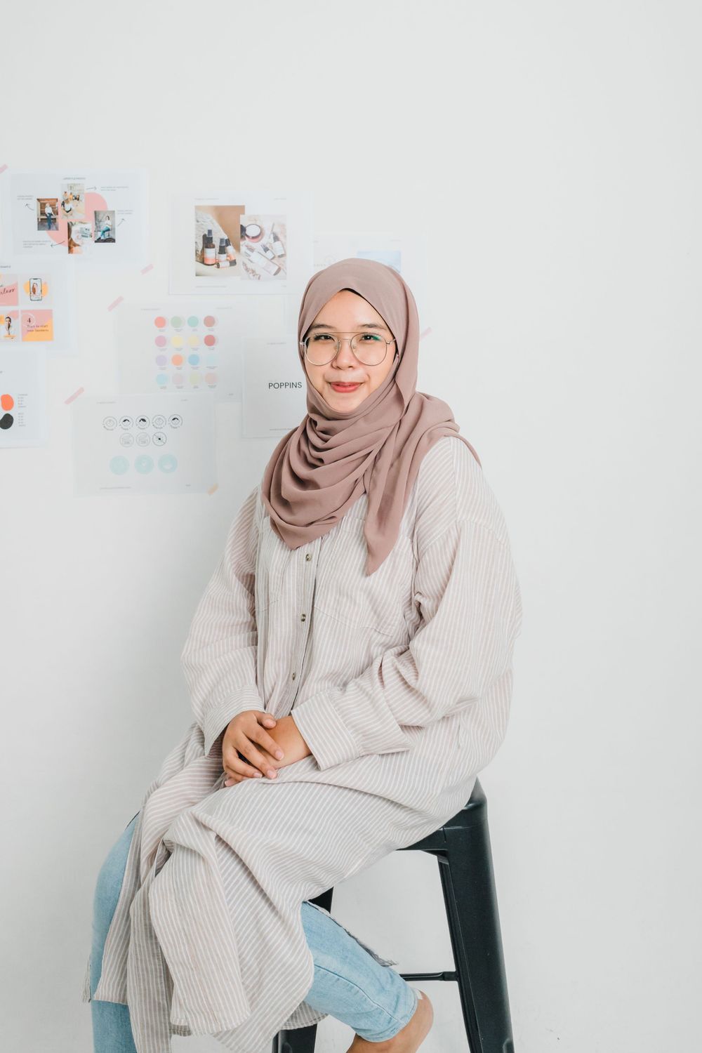 Person in light colored outfit and hijab sits casually on chair against white wall.
