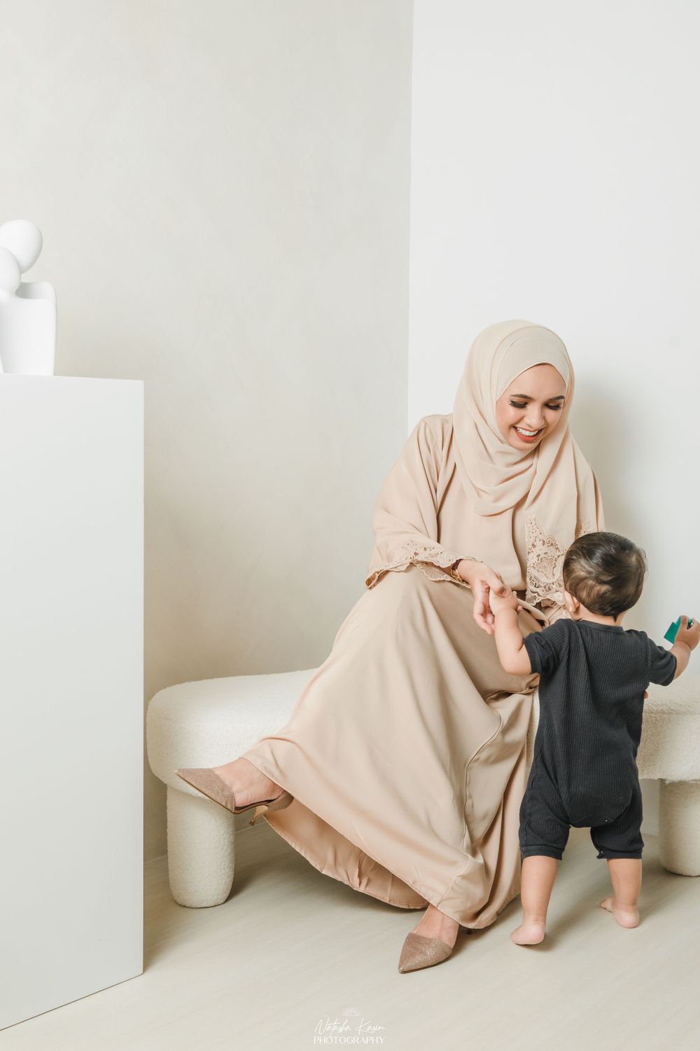 Mother in beige hijab playing with toddler on floor in minimalist white room setting.