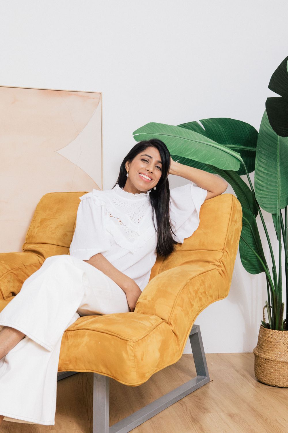 Person in white dress lounging on a yellow velvet sofa with tropical plants.
