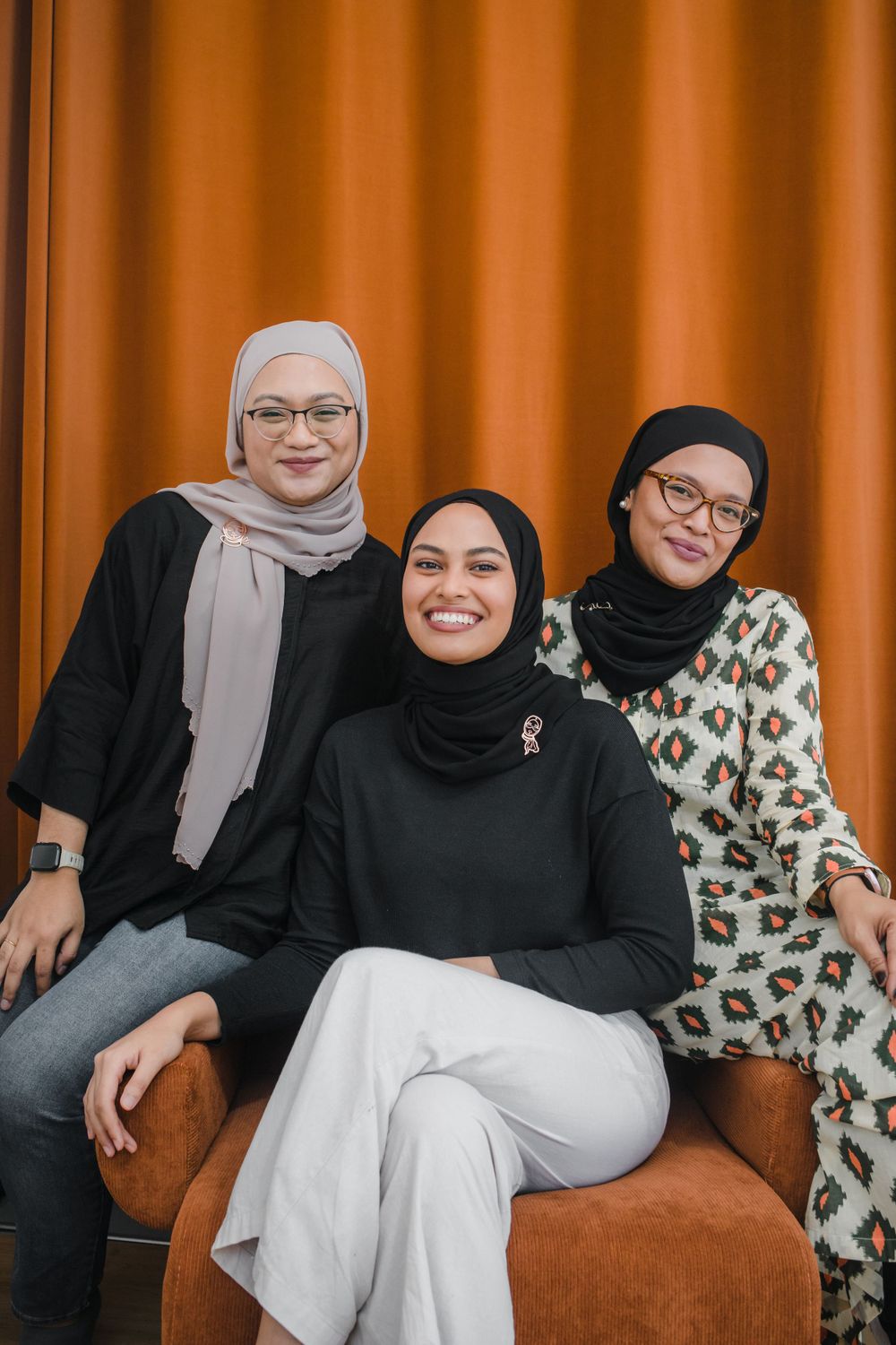 A group relaxes together on a brown couch with orange curtains in the background.