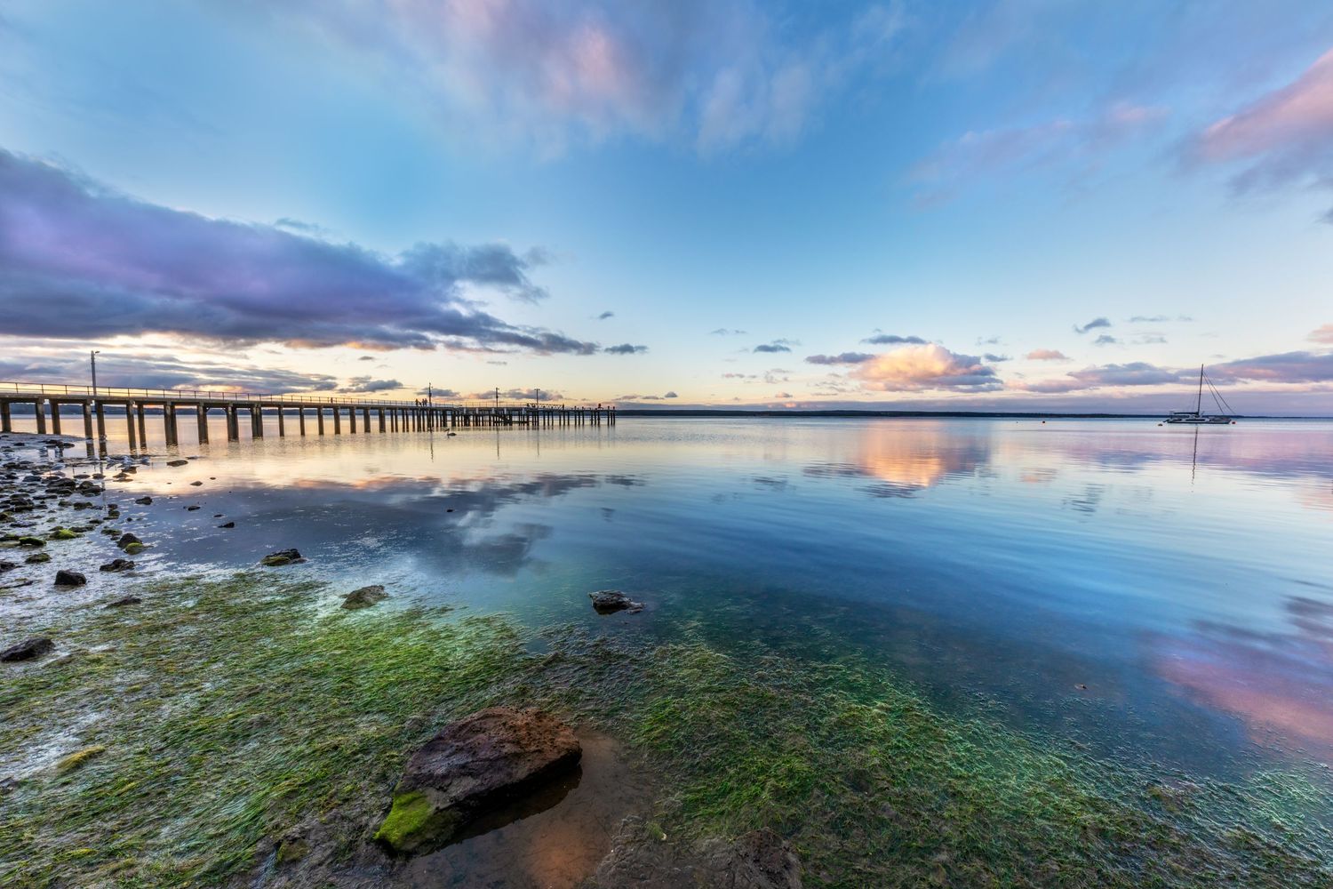 Long wooden pier stretches into calm water under pastel sunset sky.