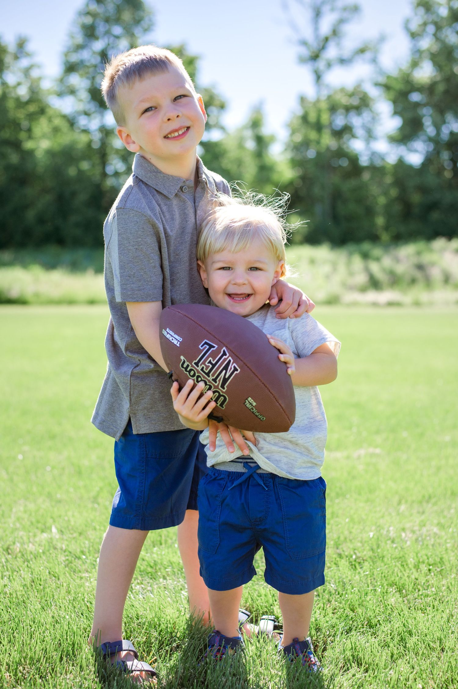 Two children pose with a football while smiling brightly in a grassy field.