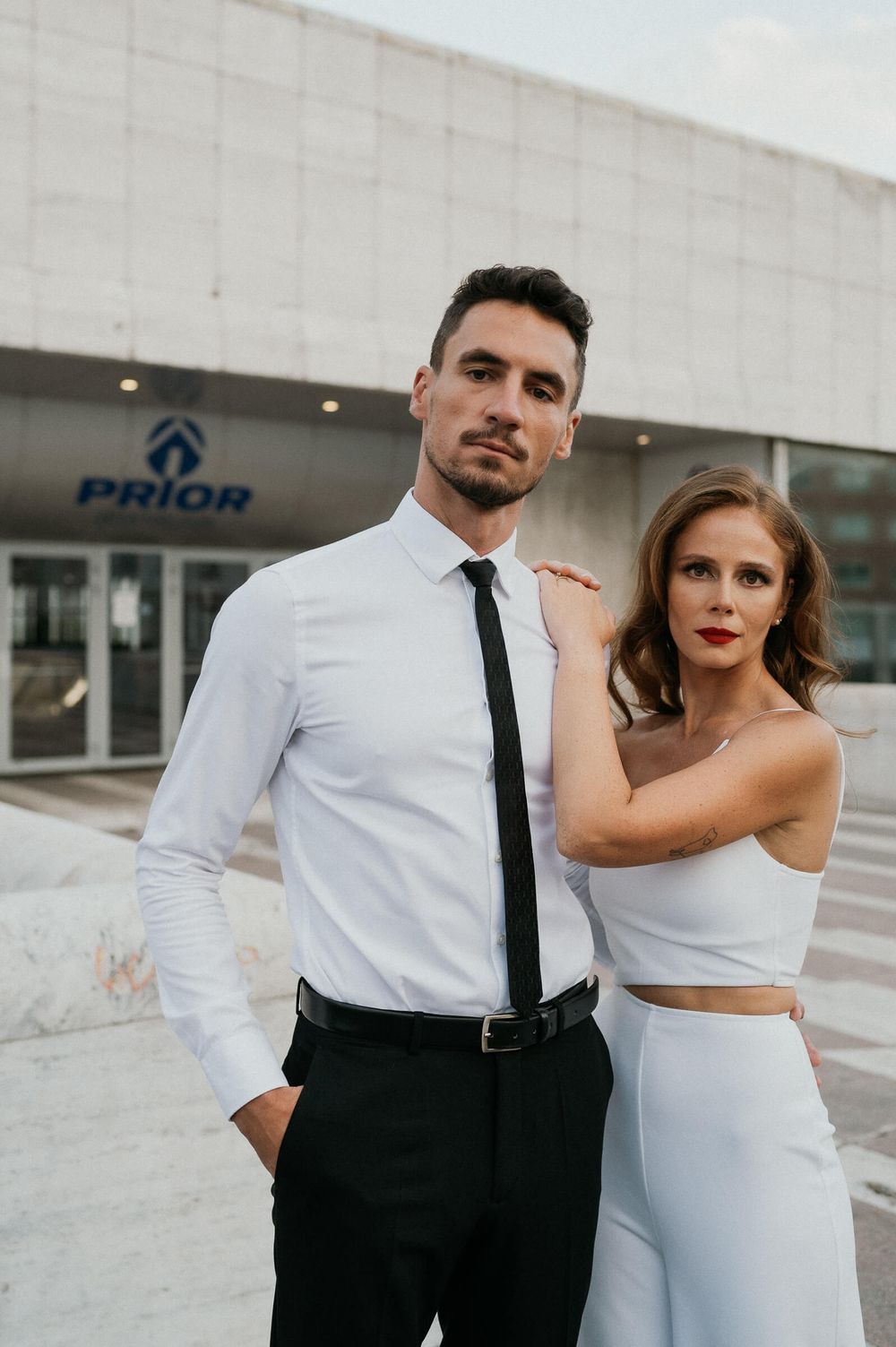 Elegant couple in formal white attire pose outside modern building.