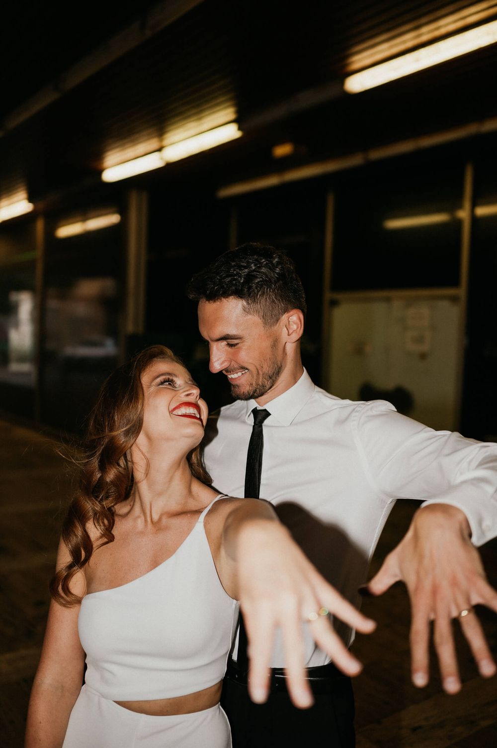 A playful dancing couple in white and black formal wear under industrial lighting.