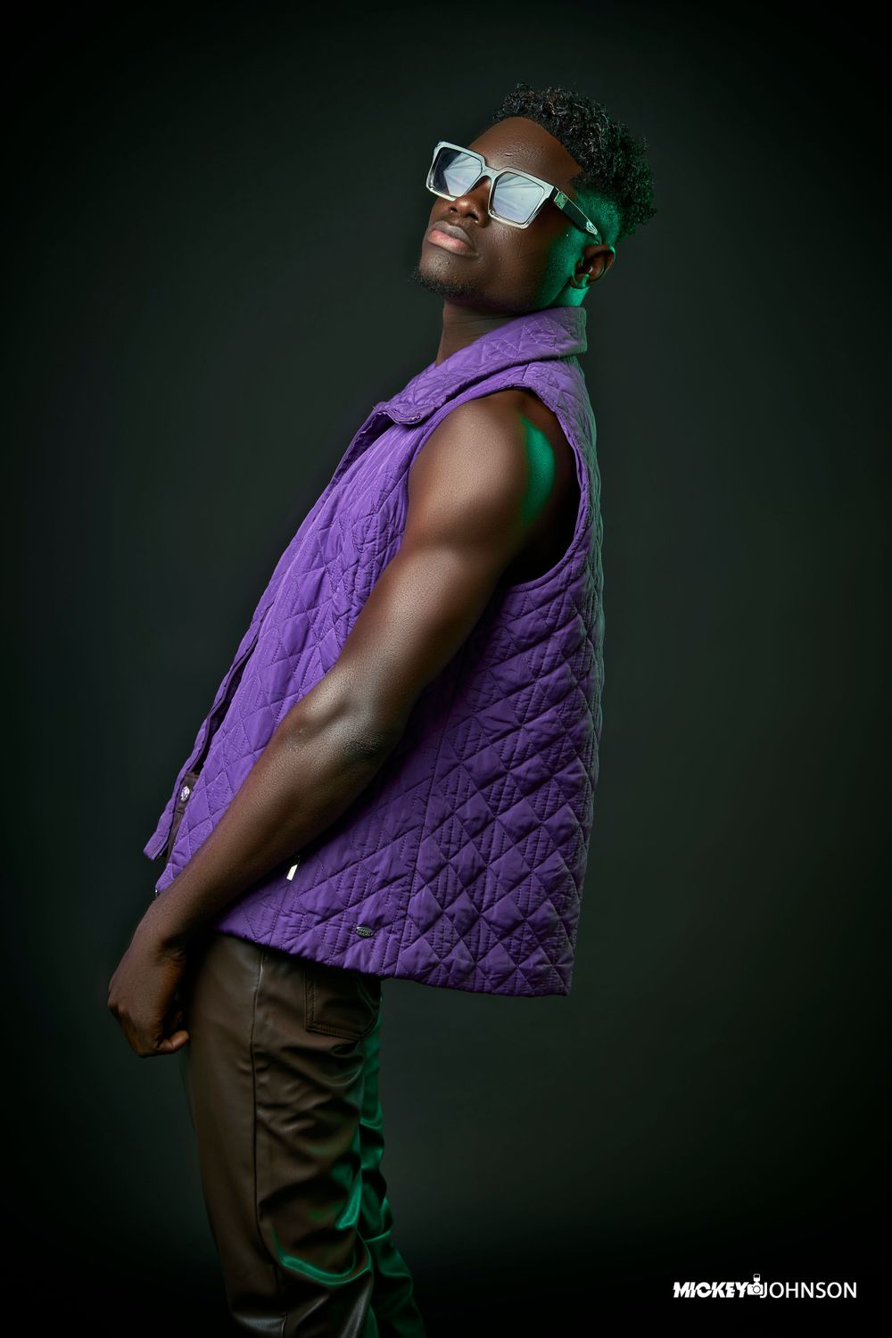 Fashion model wearing a purple quilted vest and brown pants poses against dark background.