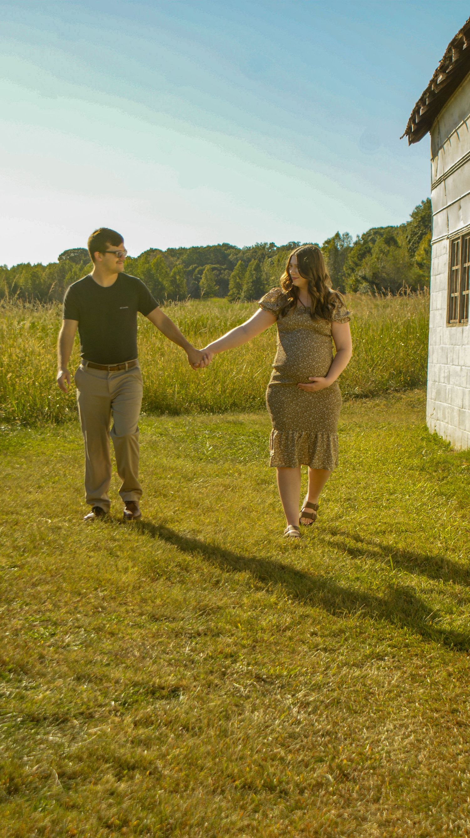 Couple walking hand in hand near a white building in a sunny field.