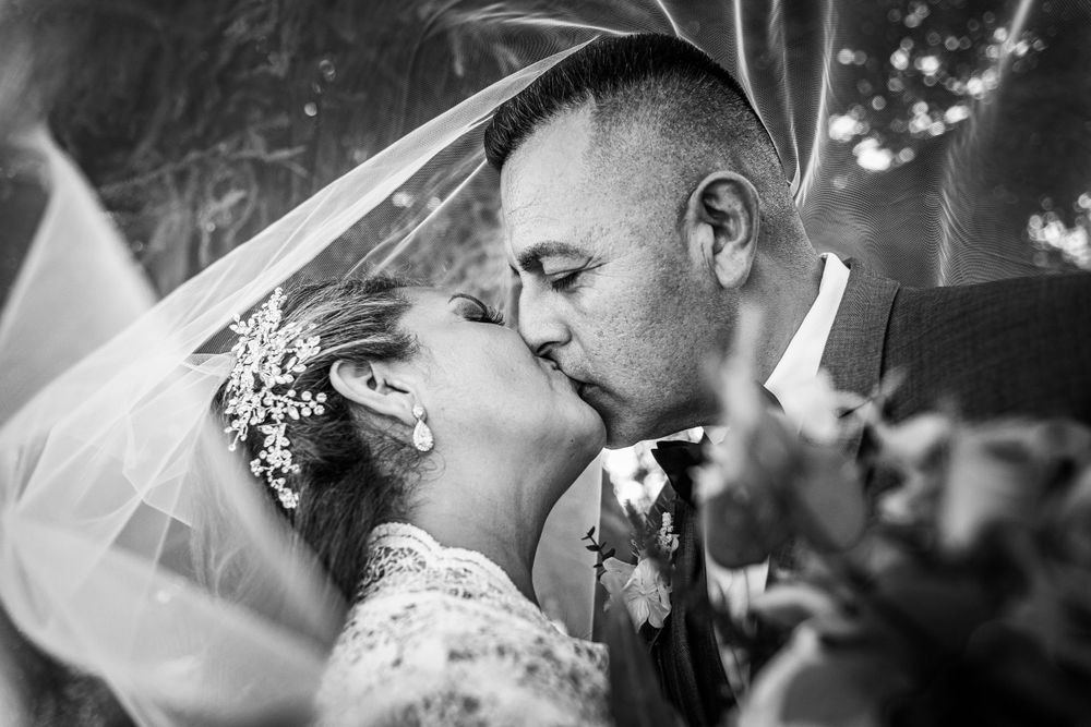 A black and white portrait of newlyweds sharing a romantic kiss under a flowing veil.