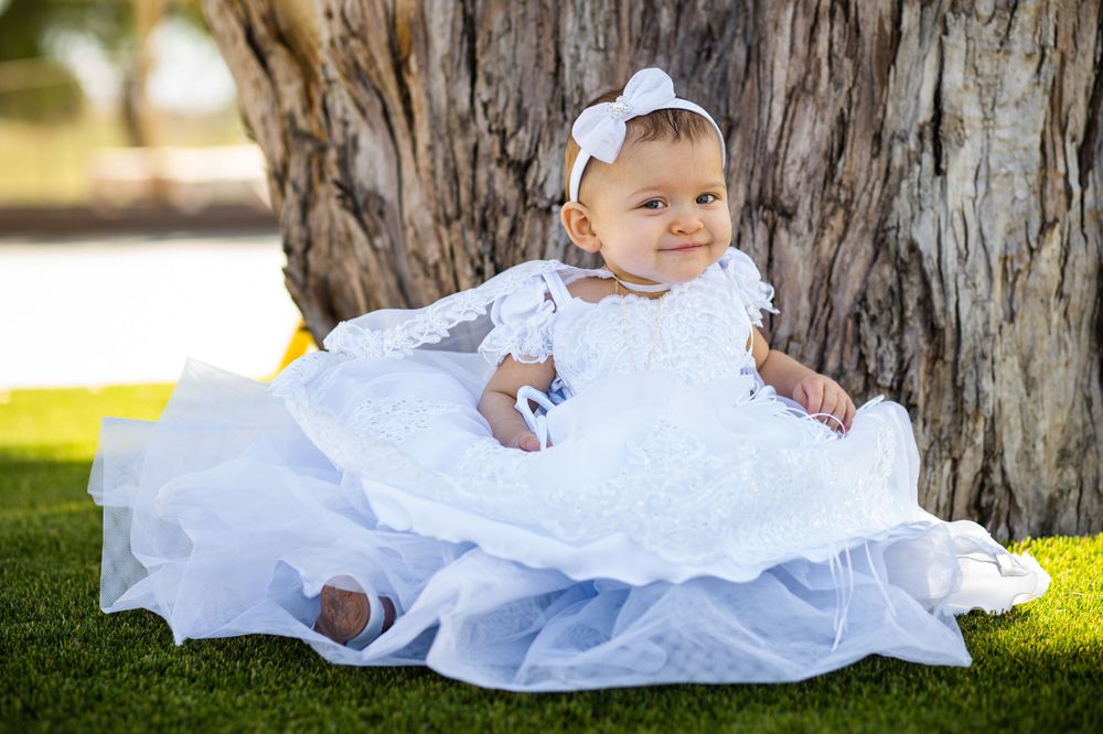 A baby in a flowing white baptismal dress sitting by a tree trunk in sunlight.
