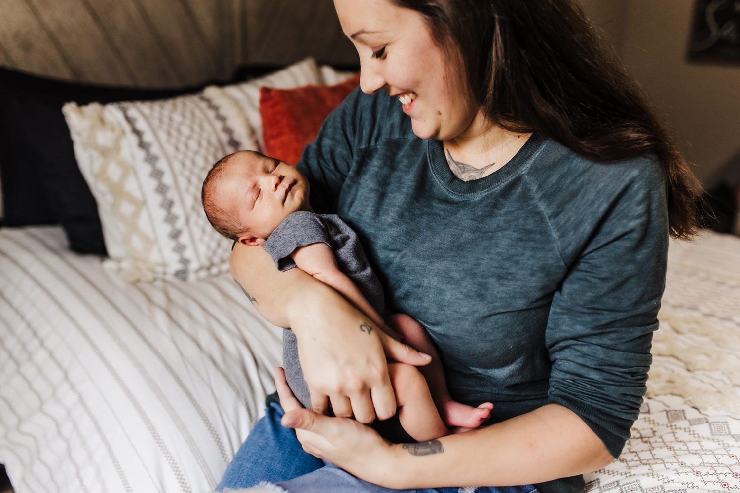 Mother holding newborn baby while sitting on bed with white and beige decor.