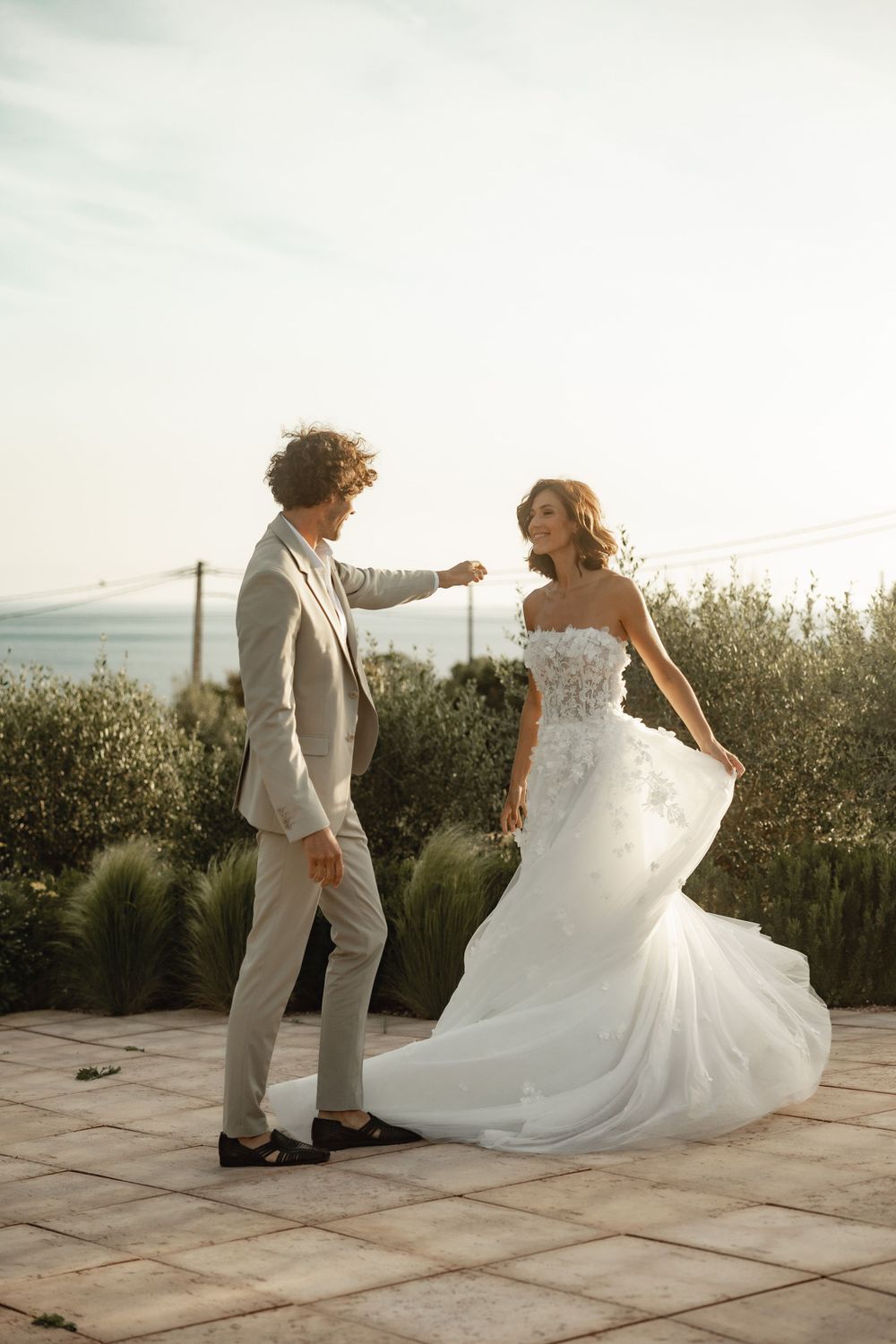 A couple in wedding attire dances playfully on a stone patio surrounded by greenery.