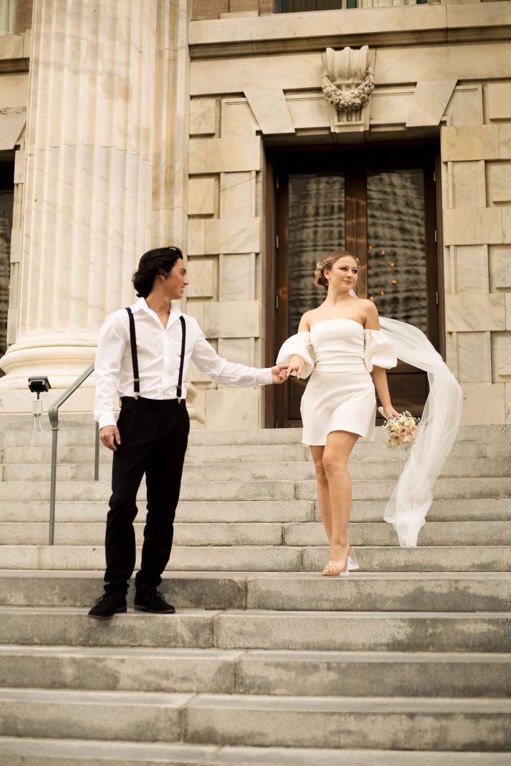 Wedding couple on grand stone steps with flowing dress and suspenders in vintage style.