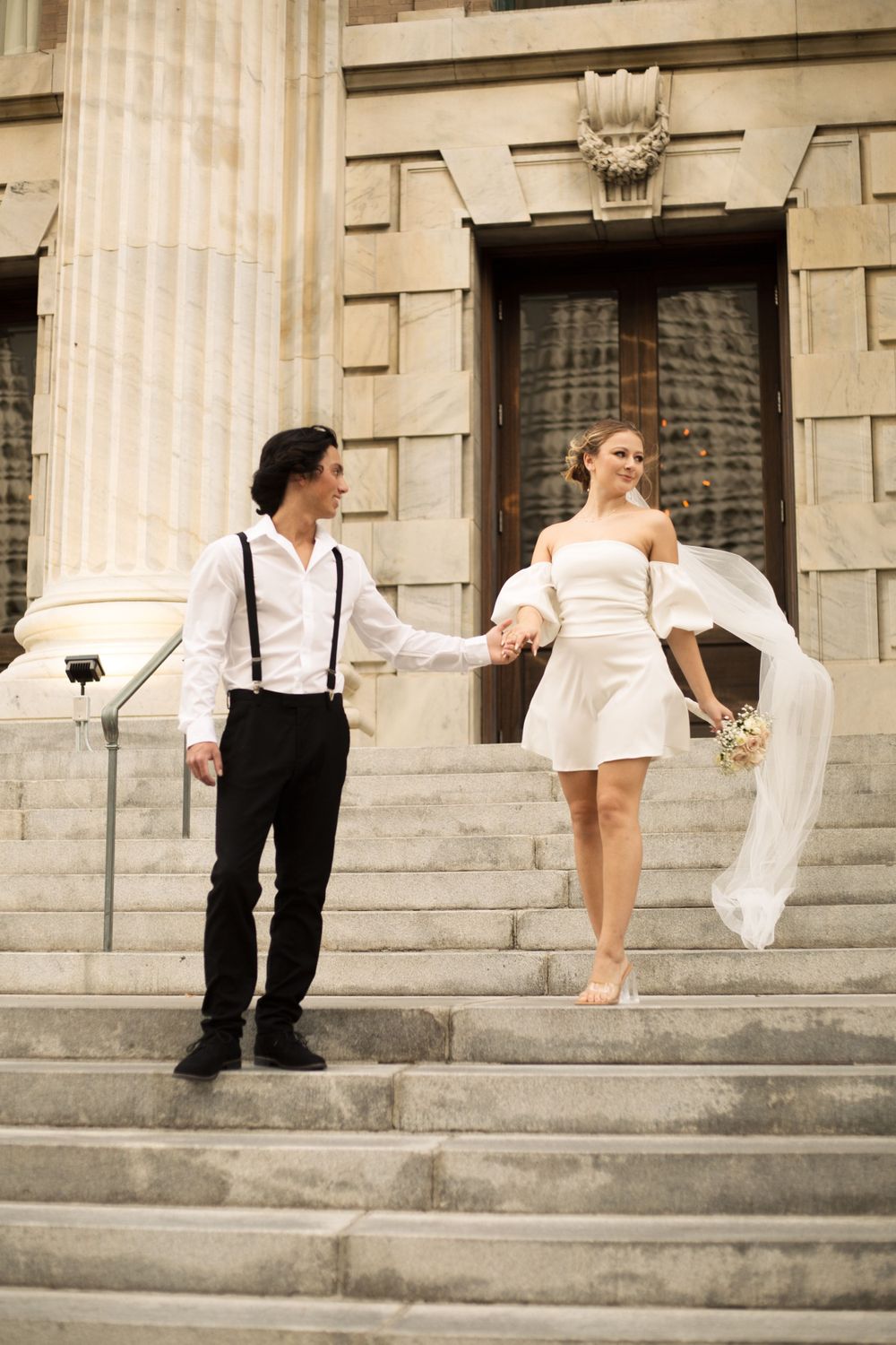 Couple in formal attire posing playfully on grand stone steps with elegant columns.