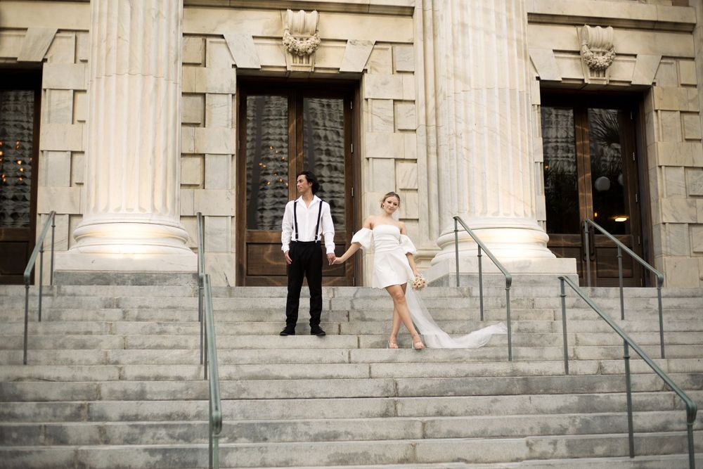 A playful wedding photo of a couple standing on stone steps with classical columns.