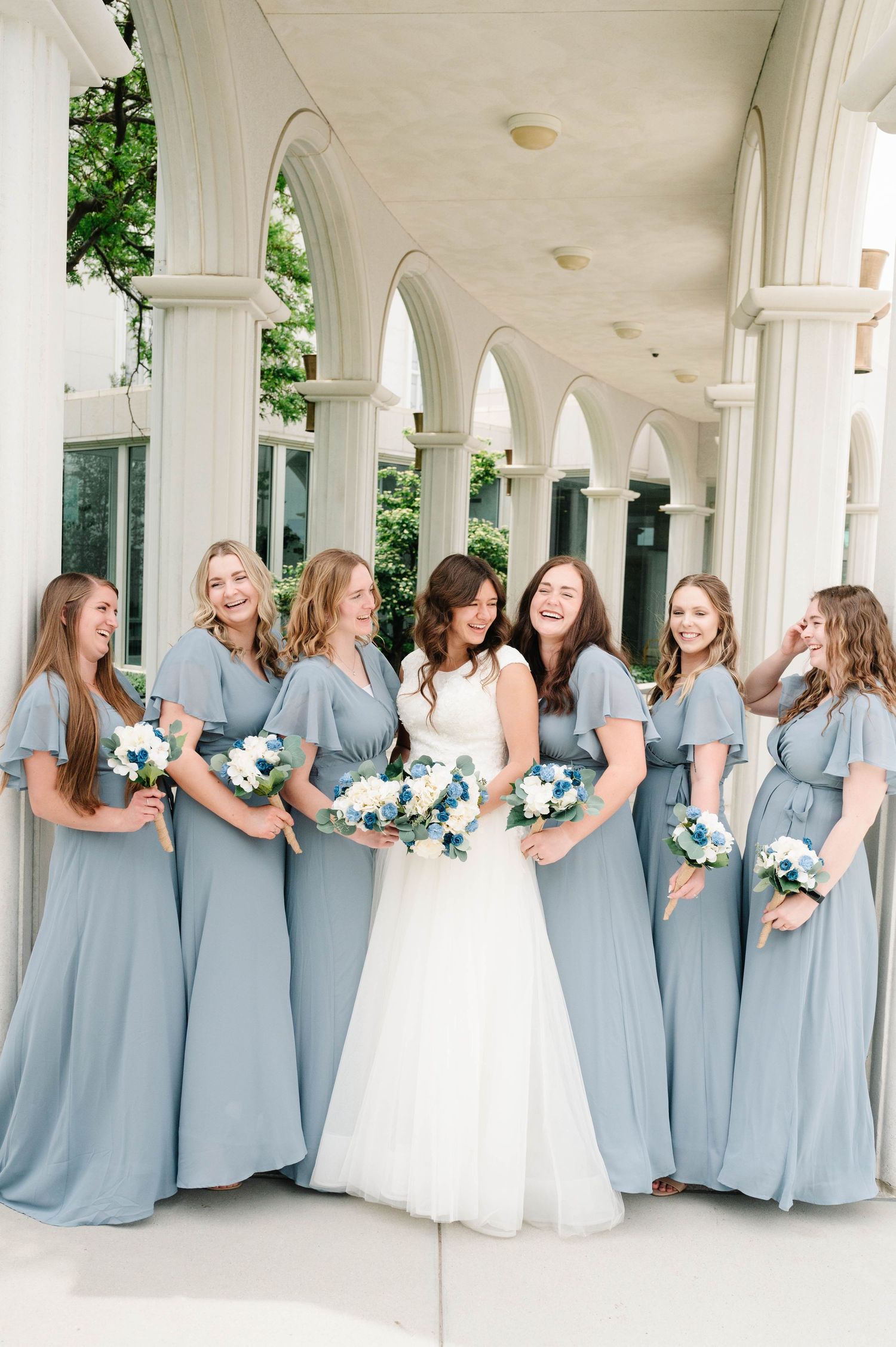 Wedding party in powder blue bridesmaid dresses posed under elegant white archway.