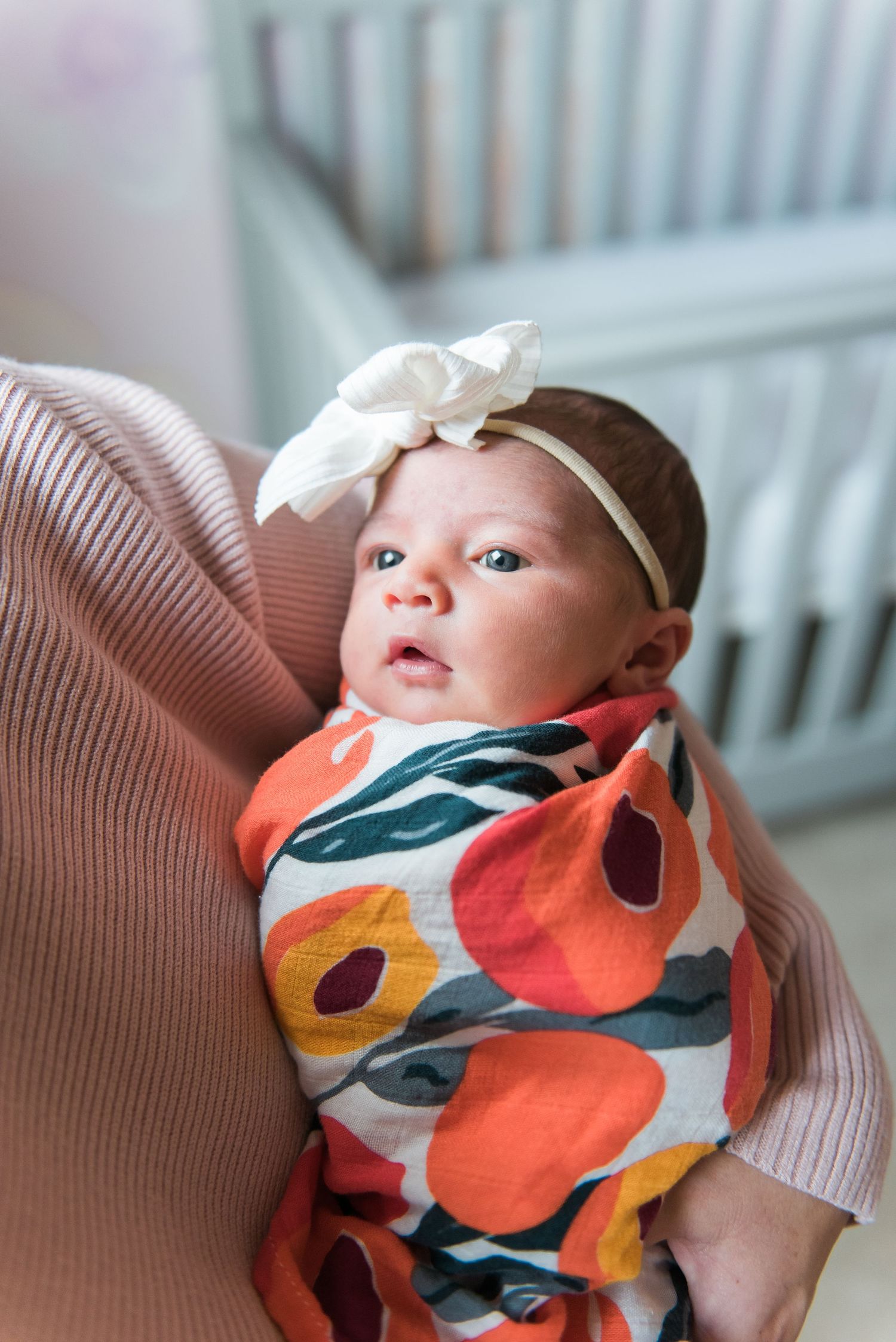 Baby wearing floral swaddle and white bow headband resting on pink cushion.