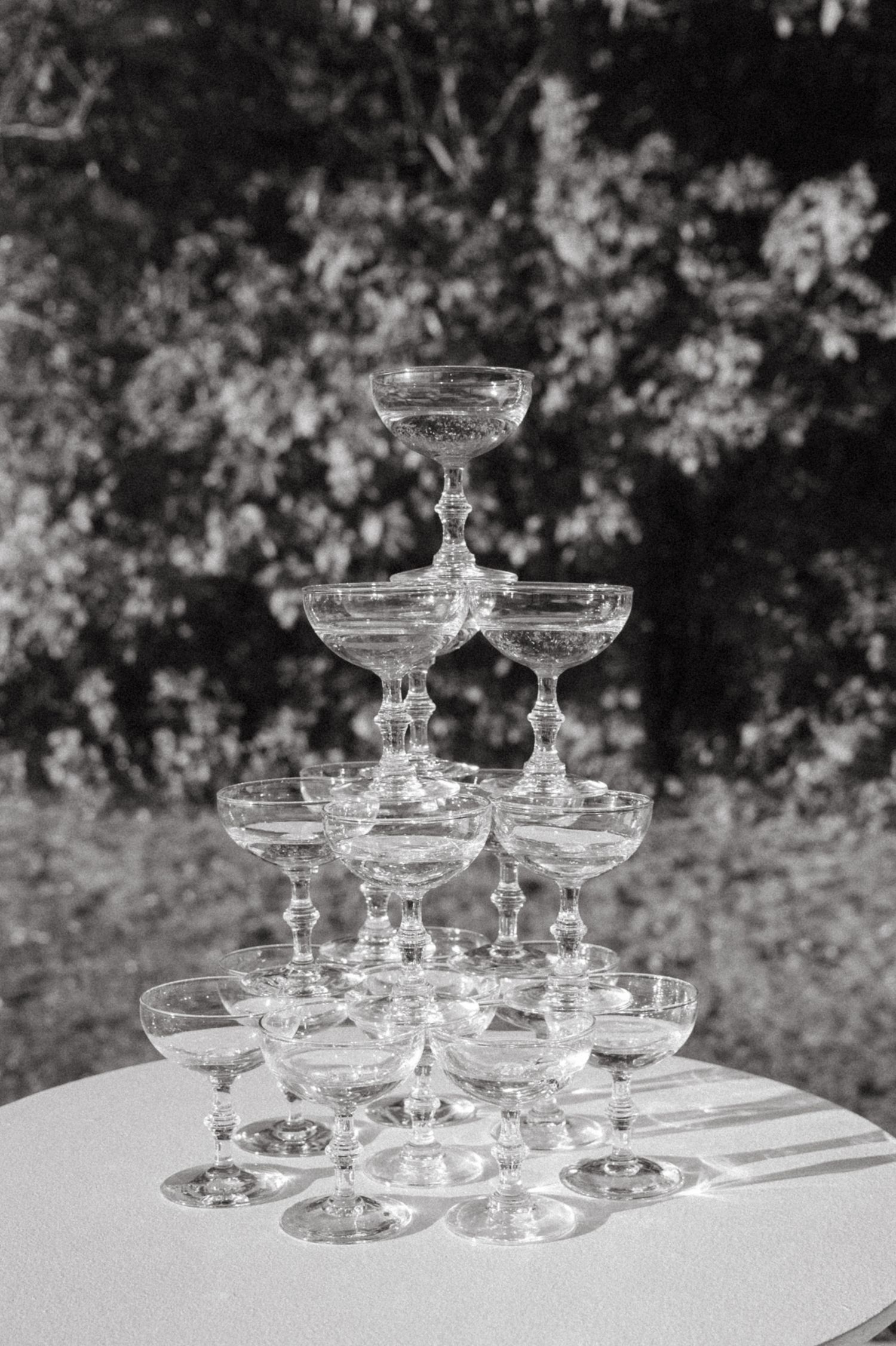 A pyramid of vintage champagne coupe glasses arranged on a white table outdoors.